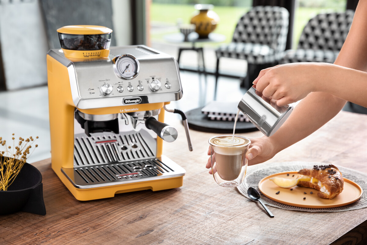 Sárga De'Longhi eszpresszógép. Egy személy tejet önt egy latte-ba. Egy croissant egy tányéron van.