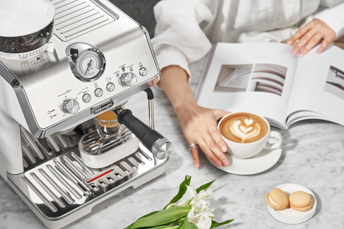 Kávéfőzőgép kávéscsészével és latte art-tal. Egy személy könyvet olvas. Macaronok és virágok az asztalon.
