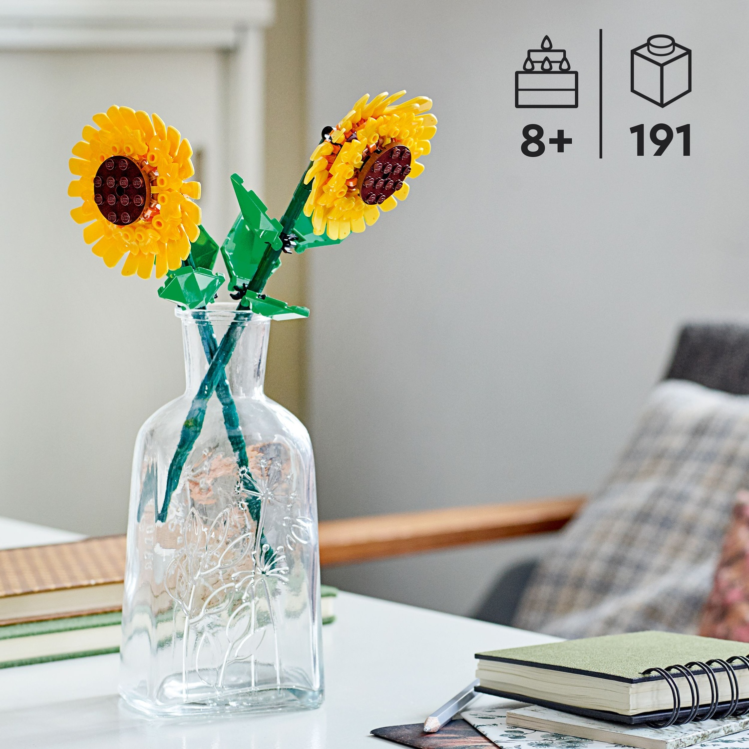 Due girasoli Lego in un vaso di vetro su un tavolo bianco. Un taccuino e una matita sono in primo piano. Lo sfondo include una sedia.