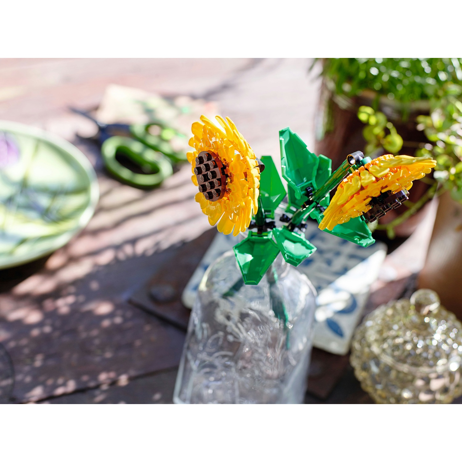 Un vaso trasparente contiene girasoli Lego. Foglie verdi, petali gialli e centri marroni. Altri oggetti sono sfocati sullo sfondo.