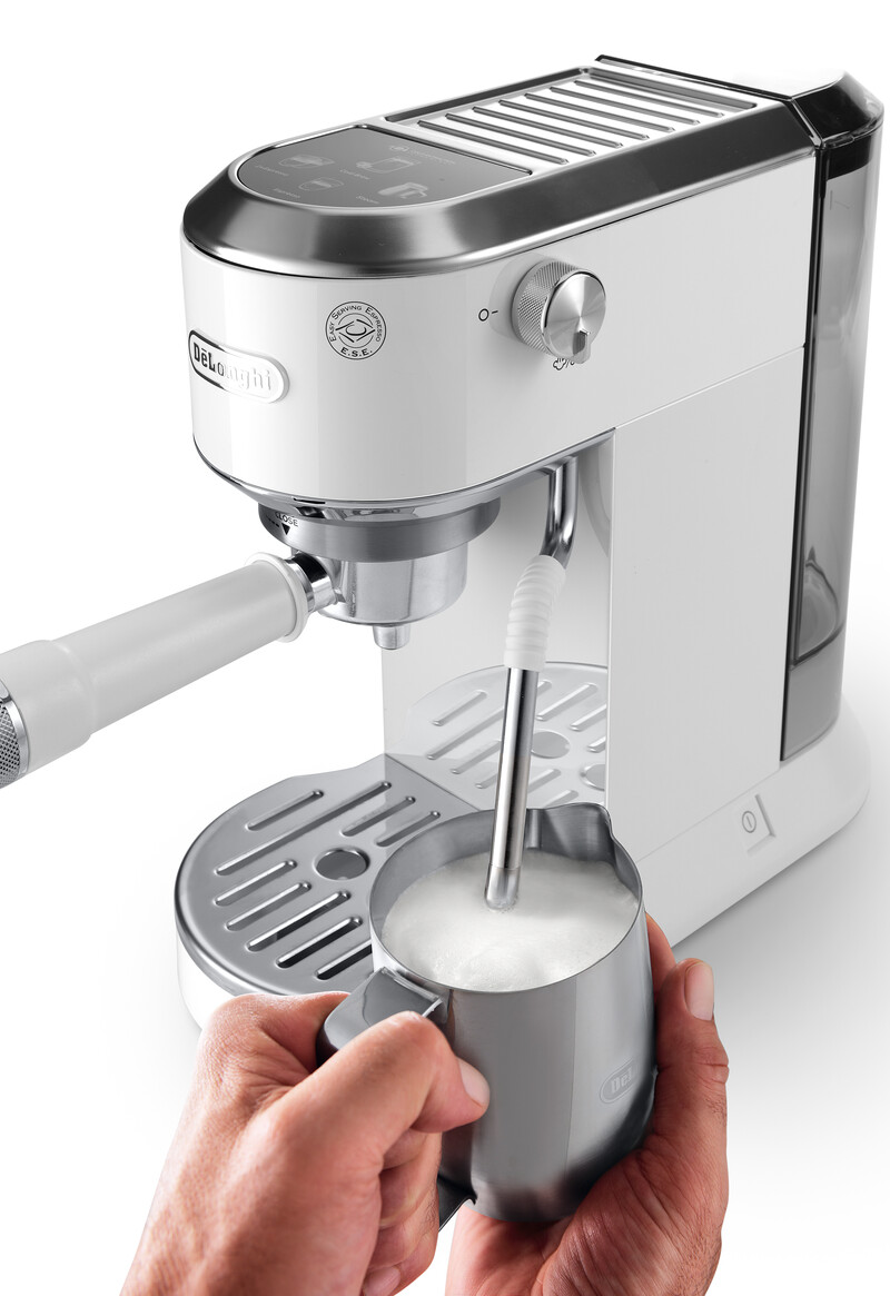 Egy fehér Delonghi eszpresszógép habosító pálcával használatban van. Egy kéz egy fémtartót tart, benne felhabosított tejjel.