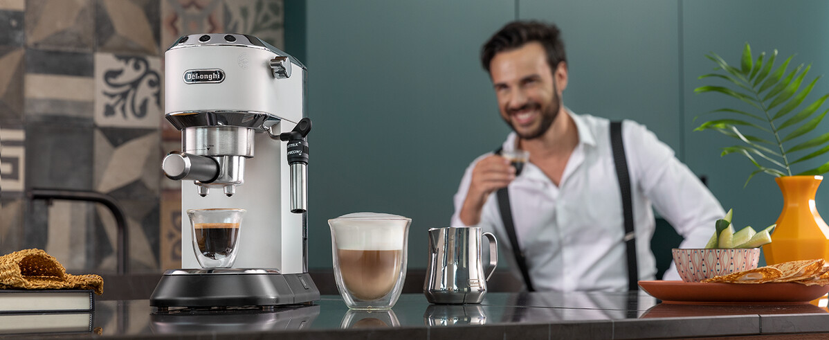 Egy fehér eszpresszógép kávéval, latte-val és egy kávéscsészét tartó, mosolygó férfival a háttérben.