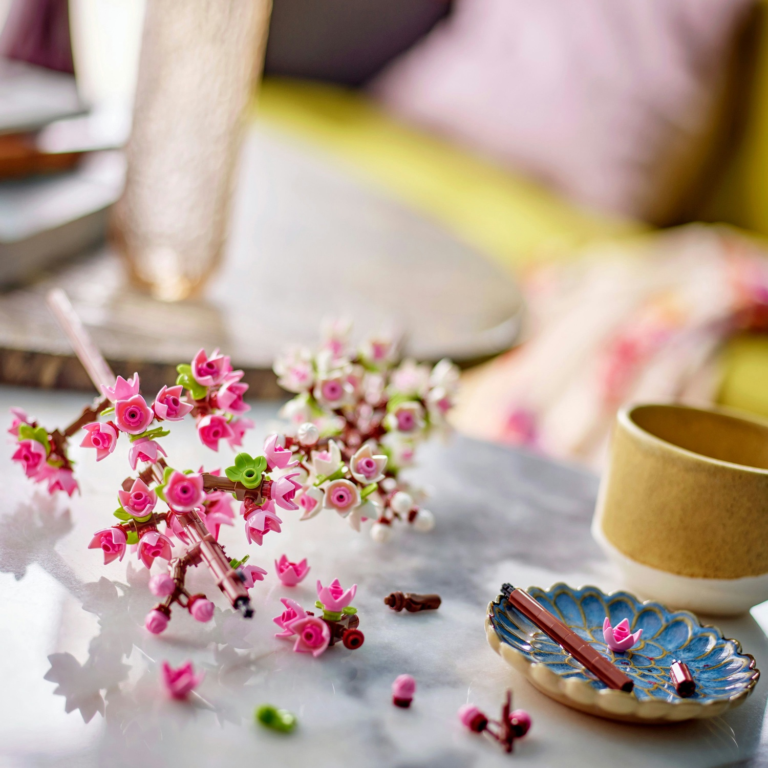 Primo piano di fiori di ciliegio Lego rosa su un tavolo di marmo. Un piatto floreale blu contiene una penna e un fiore rosa. È presente una tazza gialla.