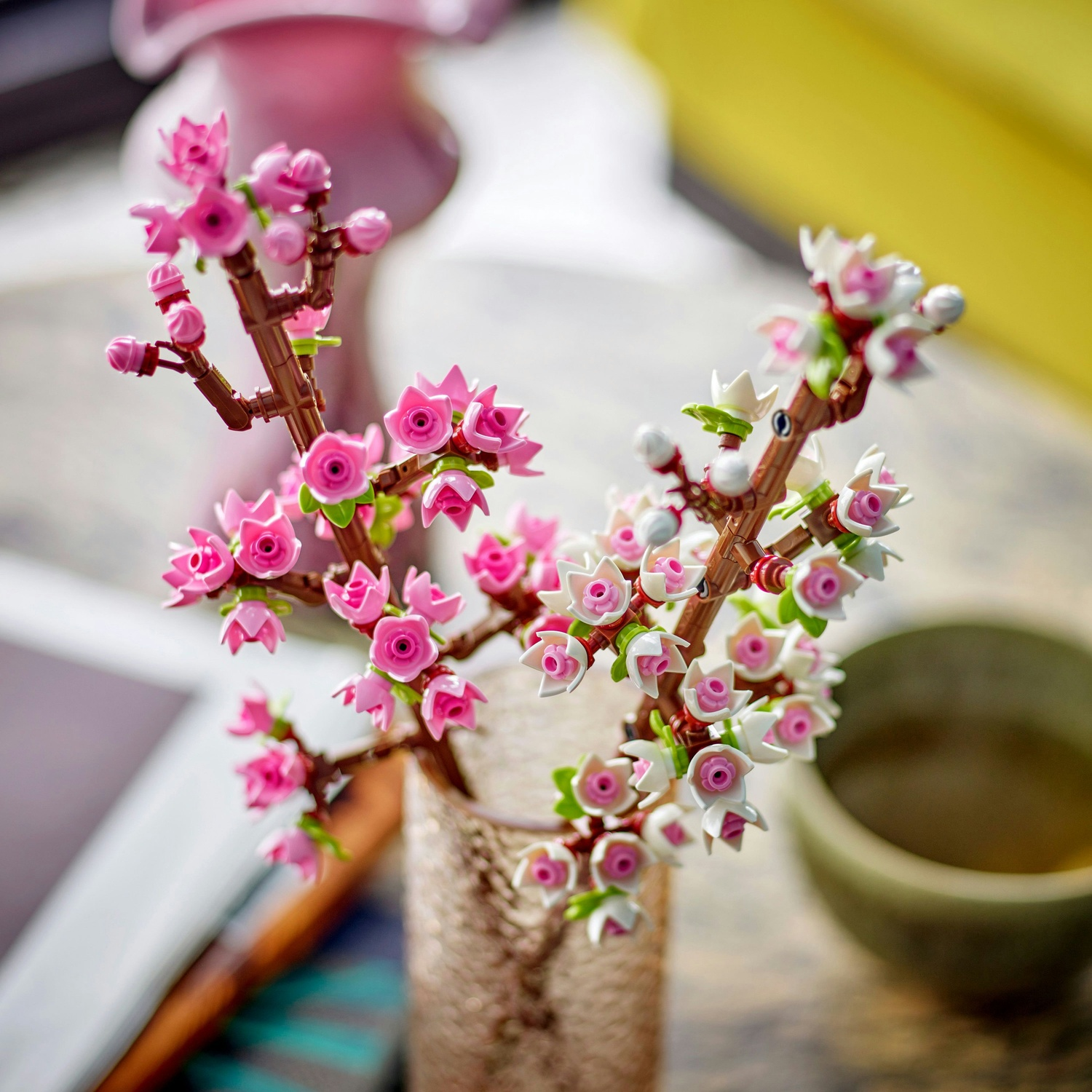 Primo piano di fiori di ciliegio in plastica rosa e bianca in un vaso di vetro. Tazza da tè verde a destra, vaso rosa sfocato dietro.