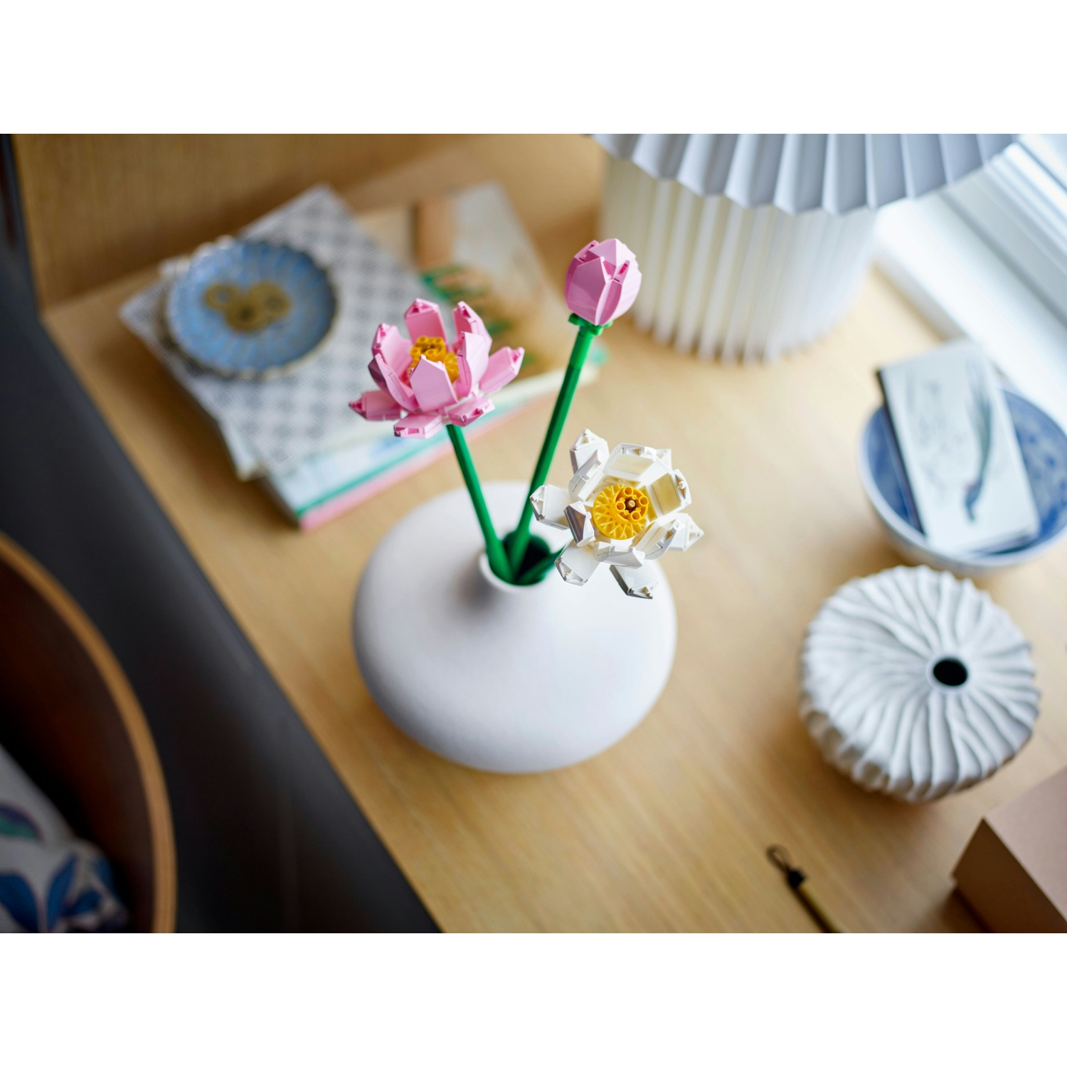 Un vaso bianco contiene fiori Lego: fiori di loto rosa e bianchi, con centri gialli e steli verdi. Su una superficie di legno, con decorazioni.