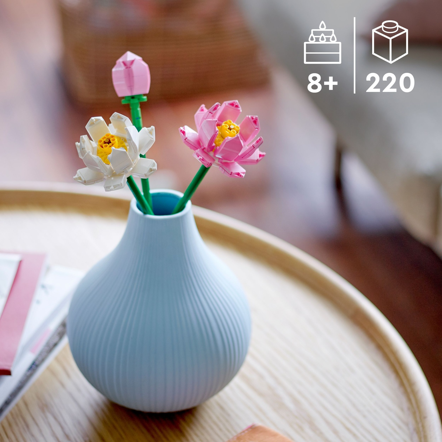 Un vaso azzurro chiaro con fiori Lego: bianchi, rosa e un bocciolo. Il vaso è su un tavolo di legno. Lo sfondo è sfocato.