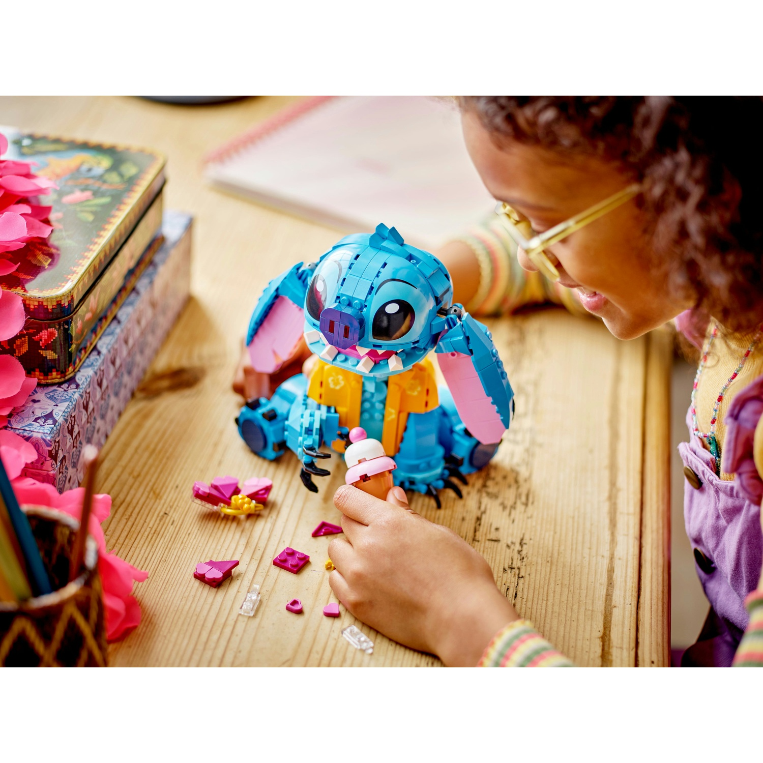 Una ragazza guarda una figura Lego di Stitch che tiene in mano un cono gelato Lego. Petali rosa e mattoncini Lego su un tavolo di legno.