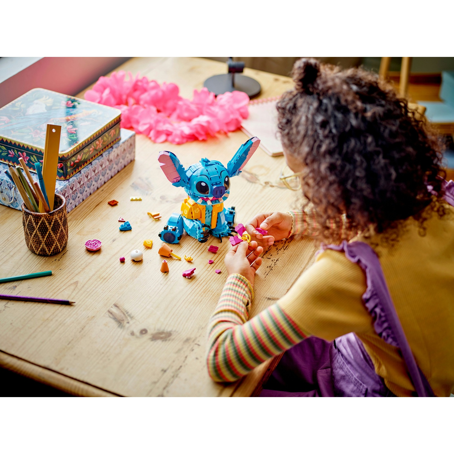 Una ragazza assembla un set Lego di Stitch blu su un tavolo di legno. Altri pezzi Lego, matite e una ghirlanda floreale rosa sono visibili.