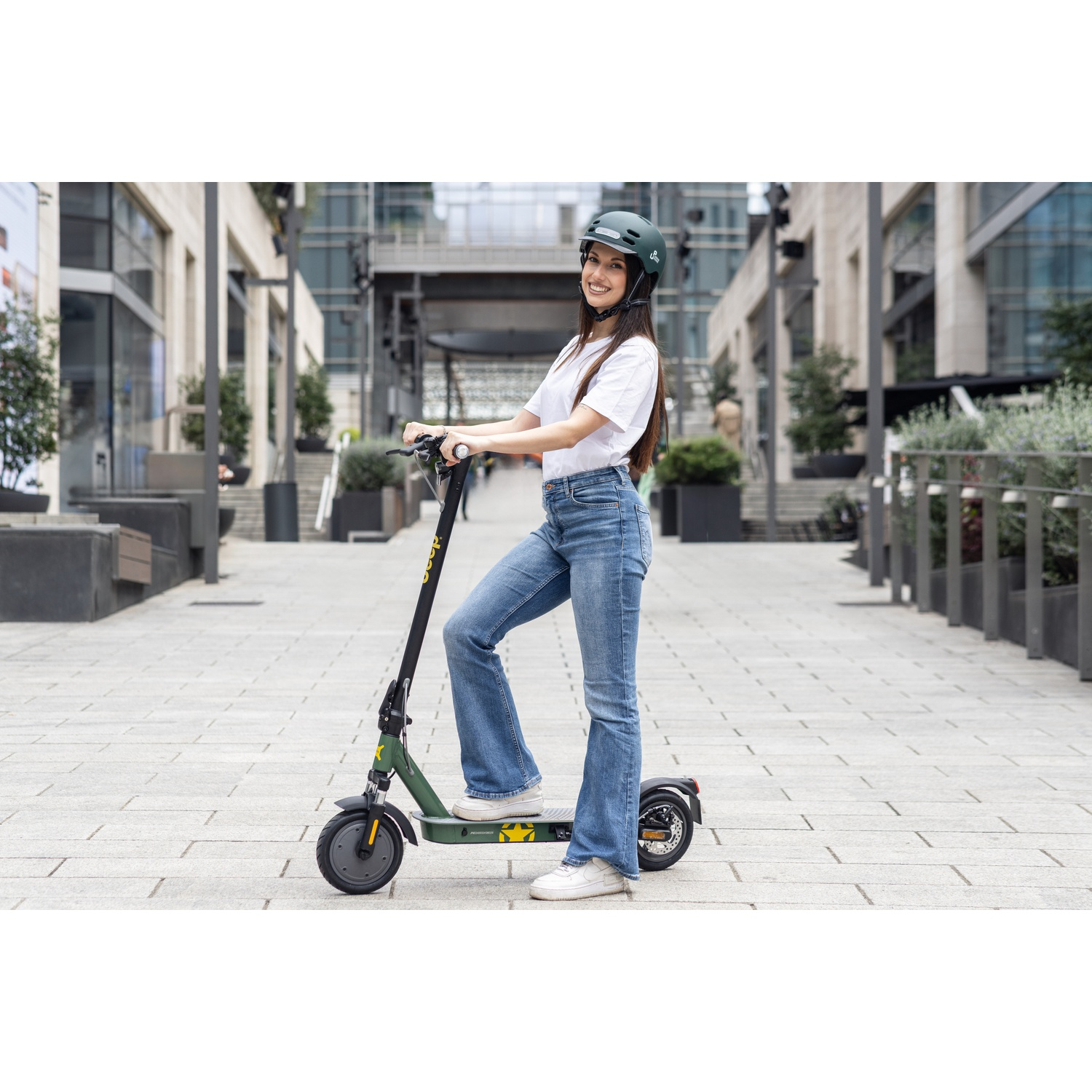 A woman smiles while standing on an e-scooter. She wears a helmet, jeans, and a white shirt. The scooter is green. Buildings are in the background.