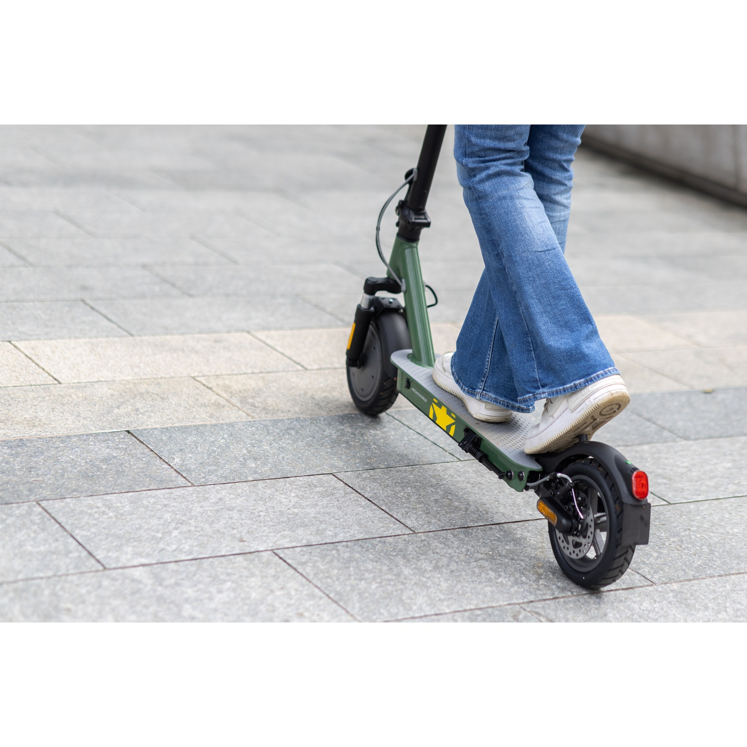 A person on a green scooter. They wear jeans and white sneakers. The scooter is on a gray paved surface.