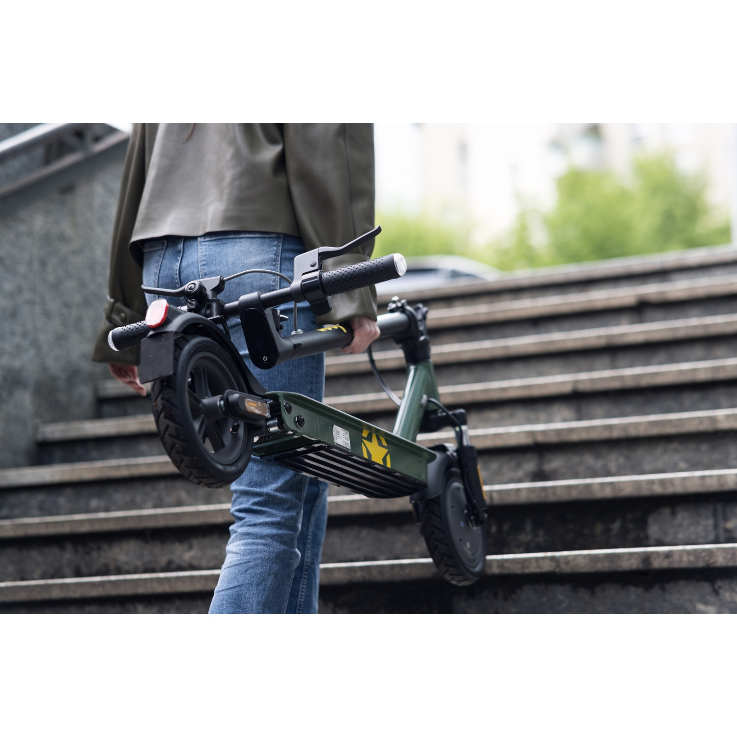 A person in jeans and jacket carries a folded green scooter up outdoor stairs. The scooter has black wheels and handlebars.