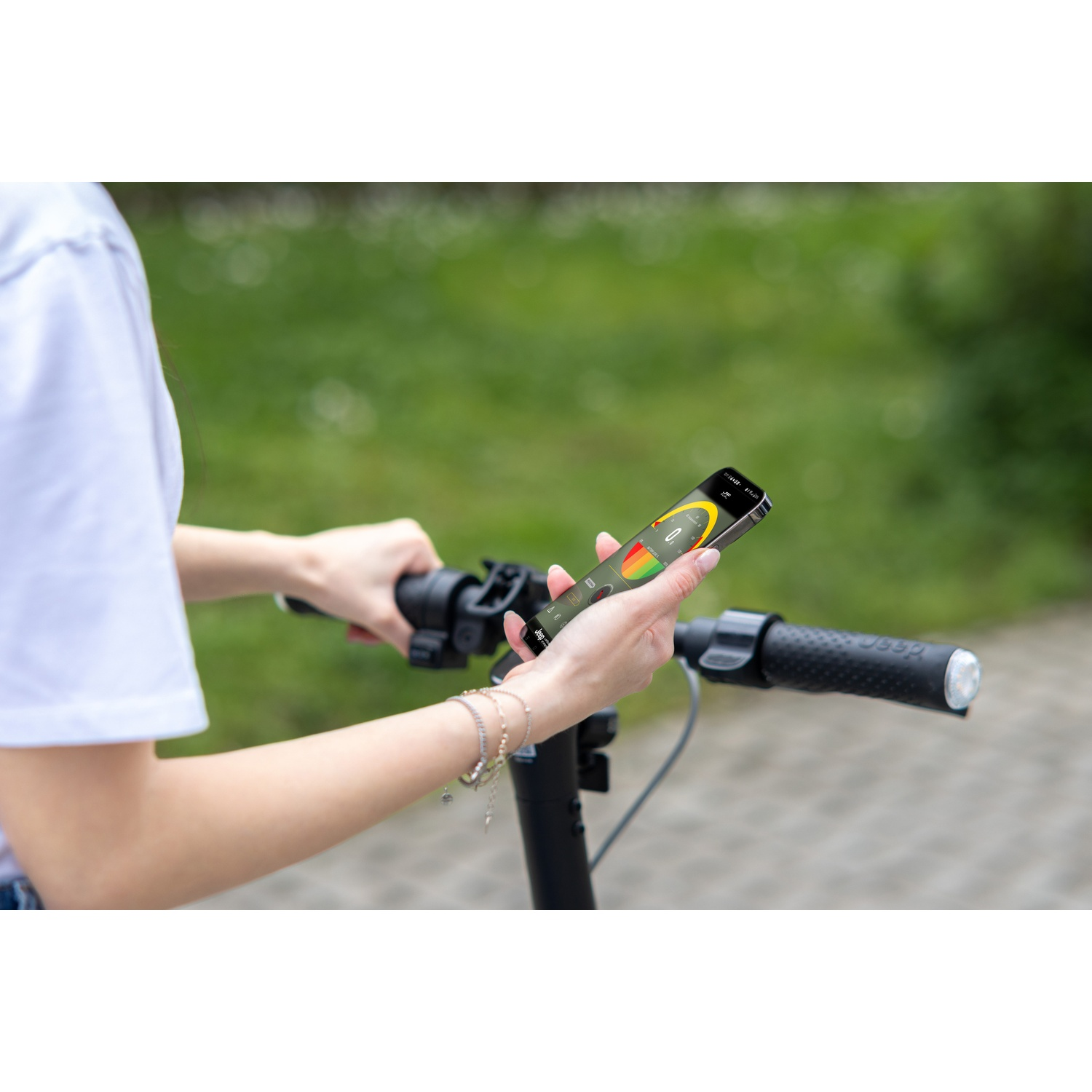 A person holds a phone displaying scooter info while holding a scooter's handlebar. Green grass in the blurry background.