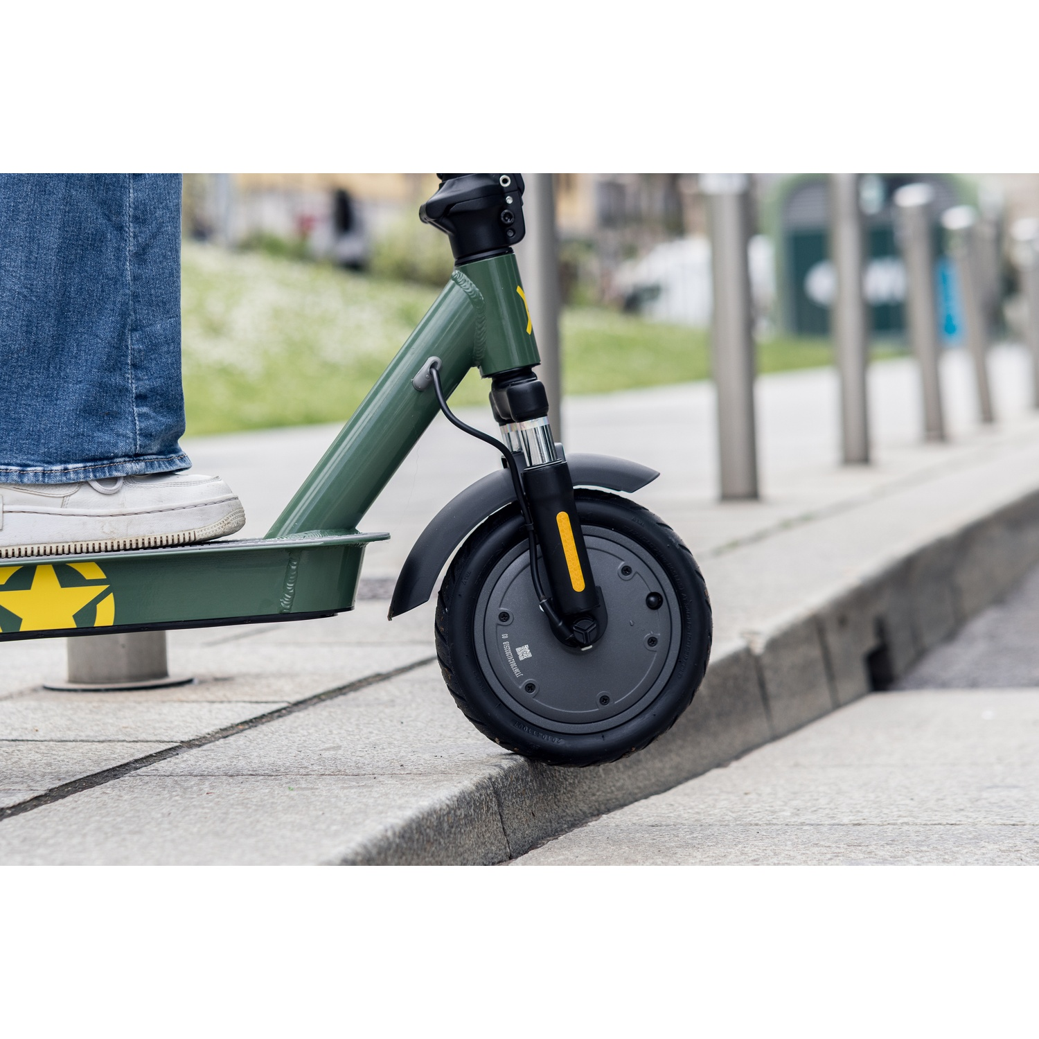 A person's foot on a green scooter. The scooter has a black wheel. The person is wearing blue jeans and white shoes.