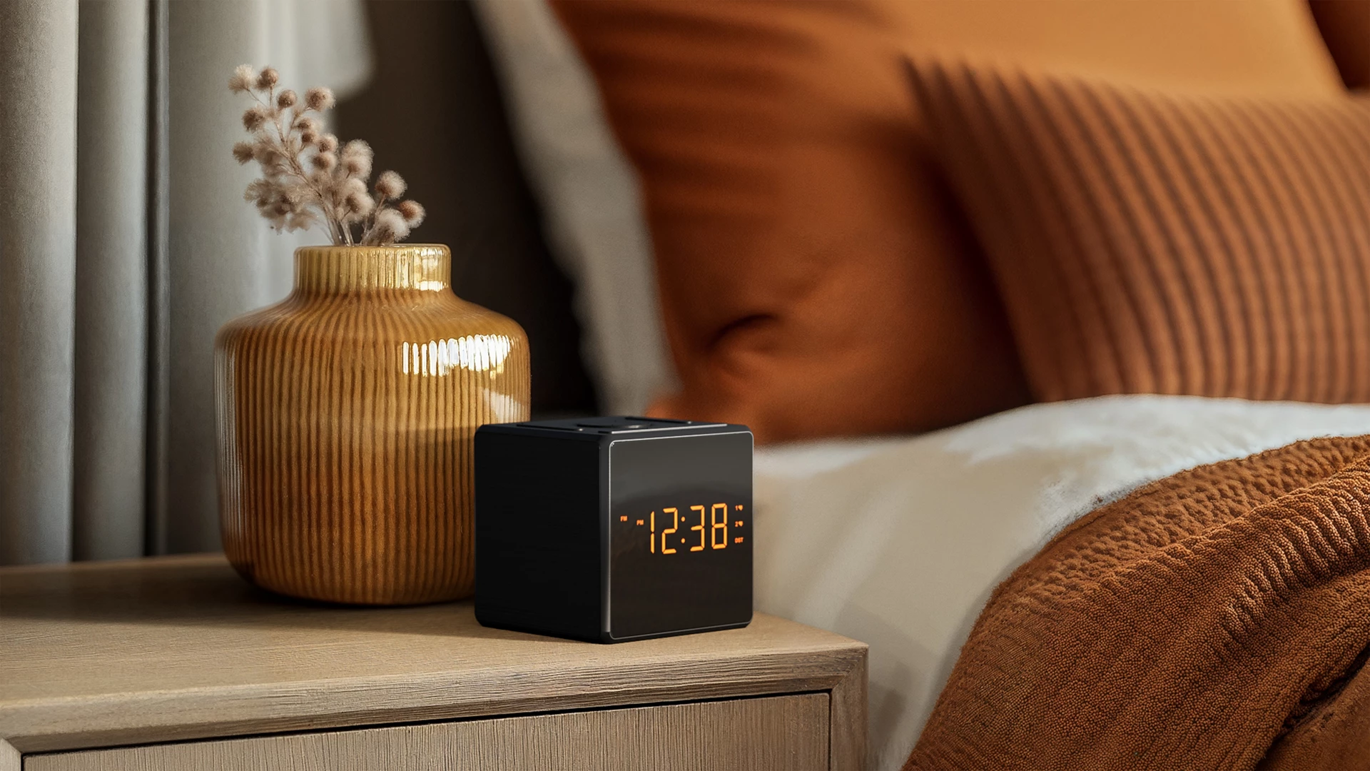 Una mesita de noche con un jarrón y un reloj. Un jarrón marrón, un reloj negro que muestra las 12:38 y una cama con almohadas naranjas.