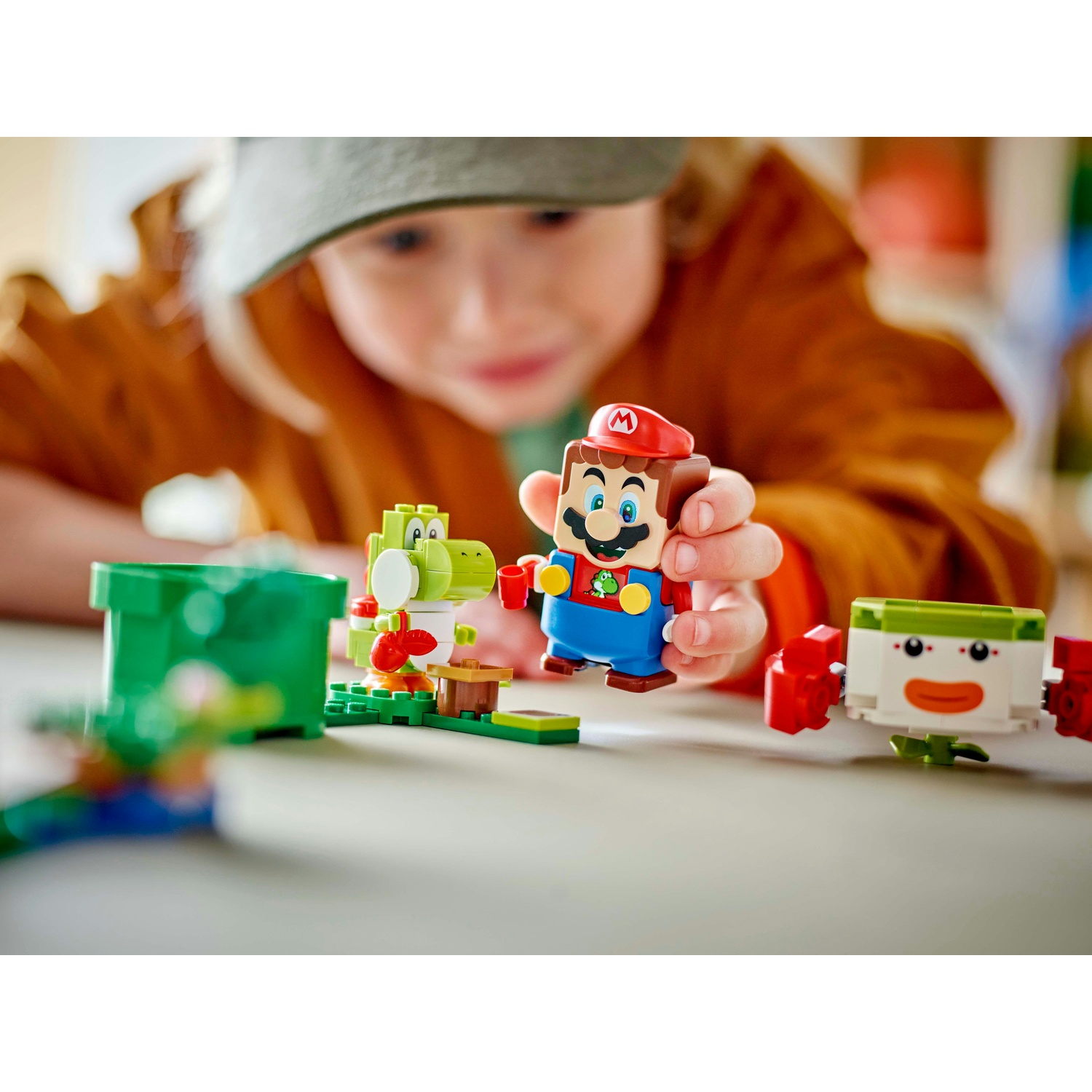 Un bambino gioca con Lego Mario, Yoshi e un personaggio. Il bambino indossa una giacca e un cappello marroni, concentrato sui giocattoli.