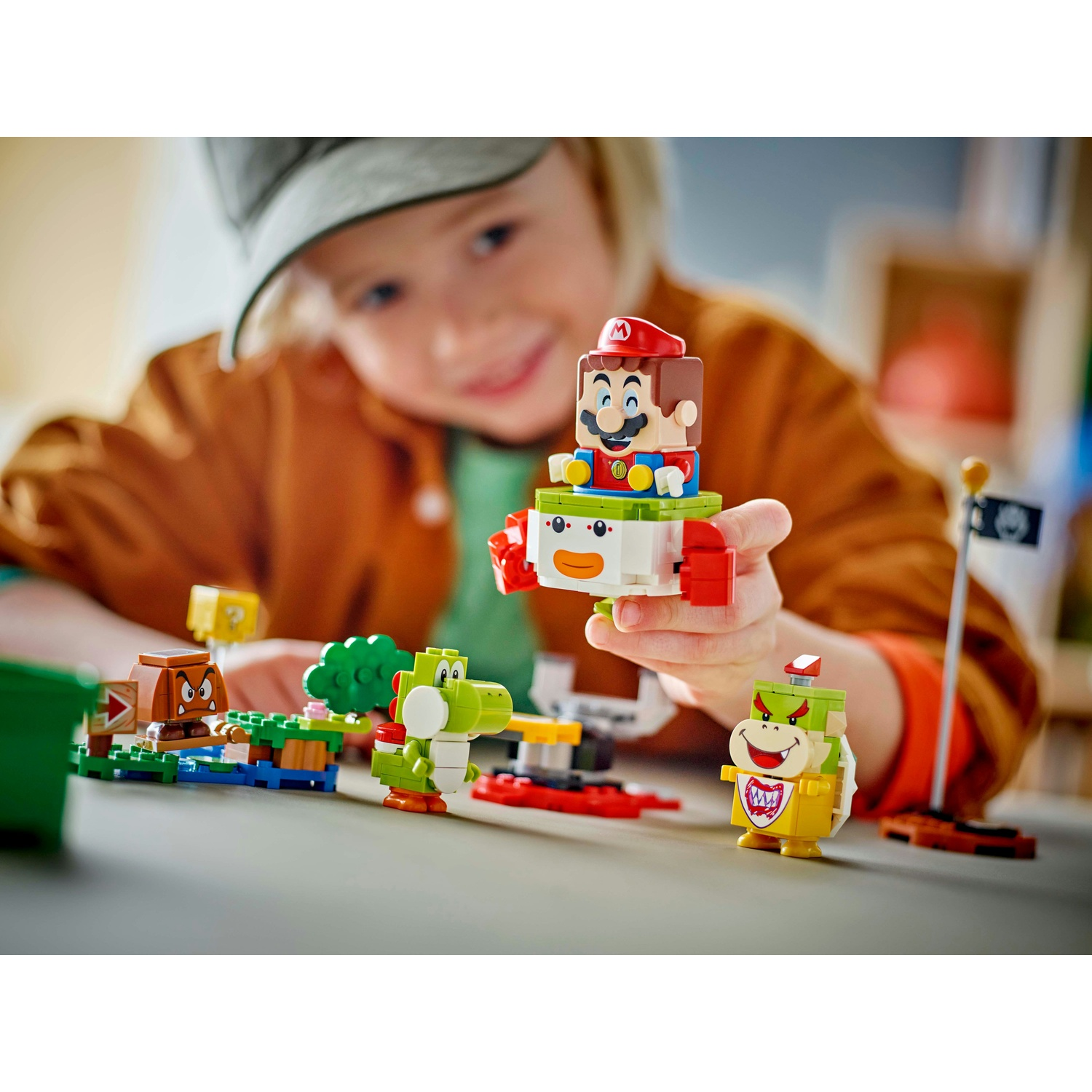 Un bambino tiene una figura LEGO di Mario. LEGO Bowser Jr. e Yoshi sono sul tavolo. Sono presenti anche un'asta portabandiera e altri elementi LEGO.