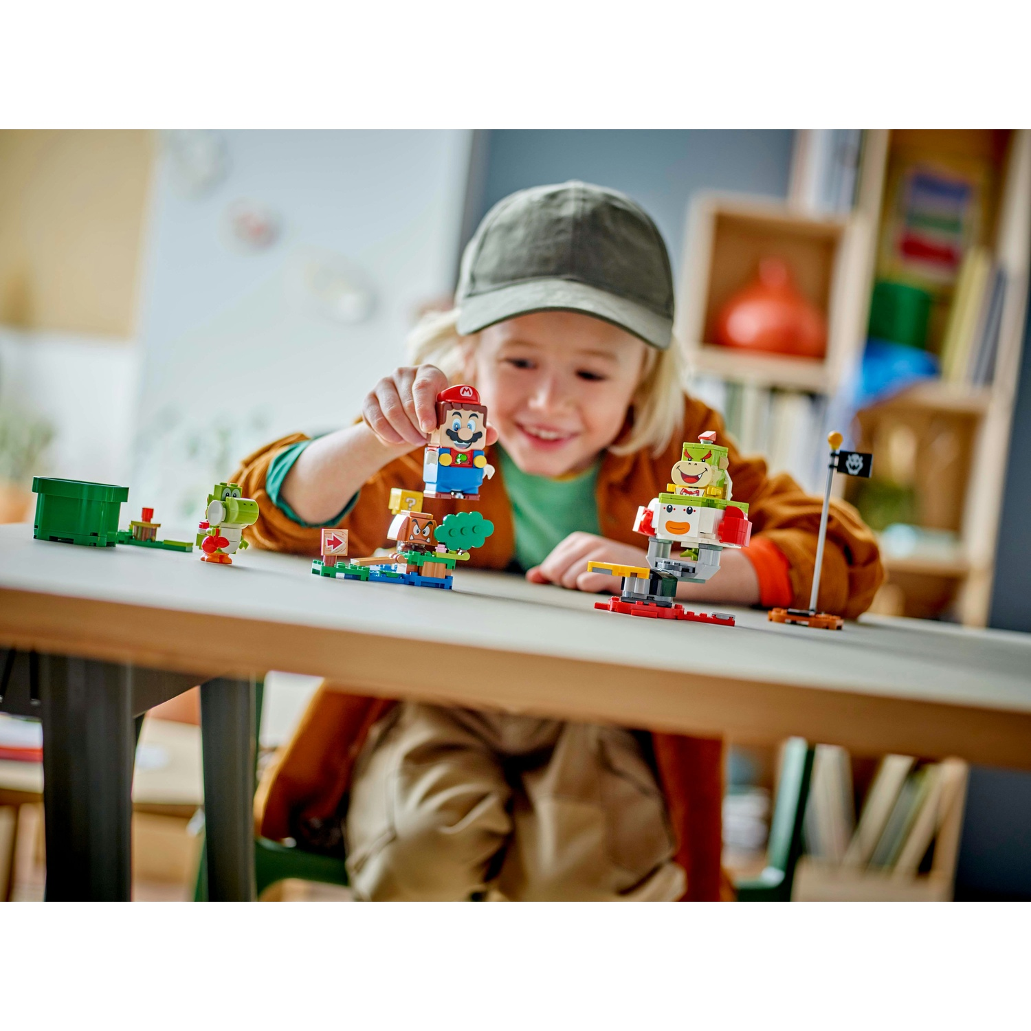 Un bambino gioca con i set Lego di Mario. Mario, Yoshi e altri personaggi sono su un tavolo. Il bambino sorride, indossa un berretto e una giacca marrone.