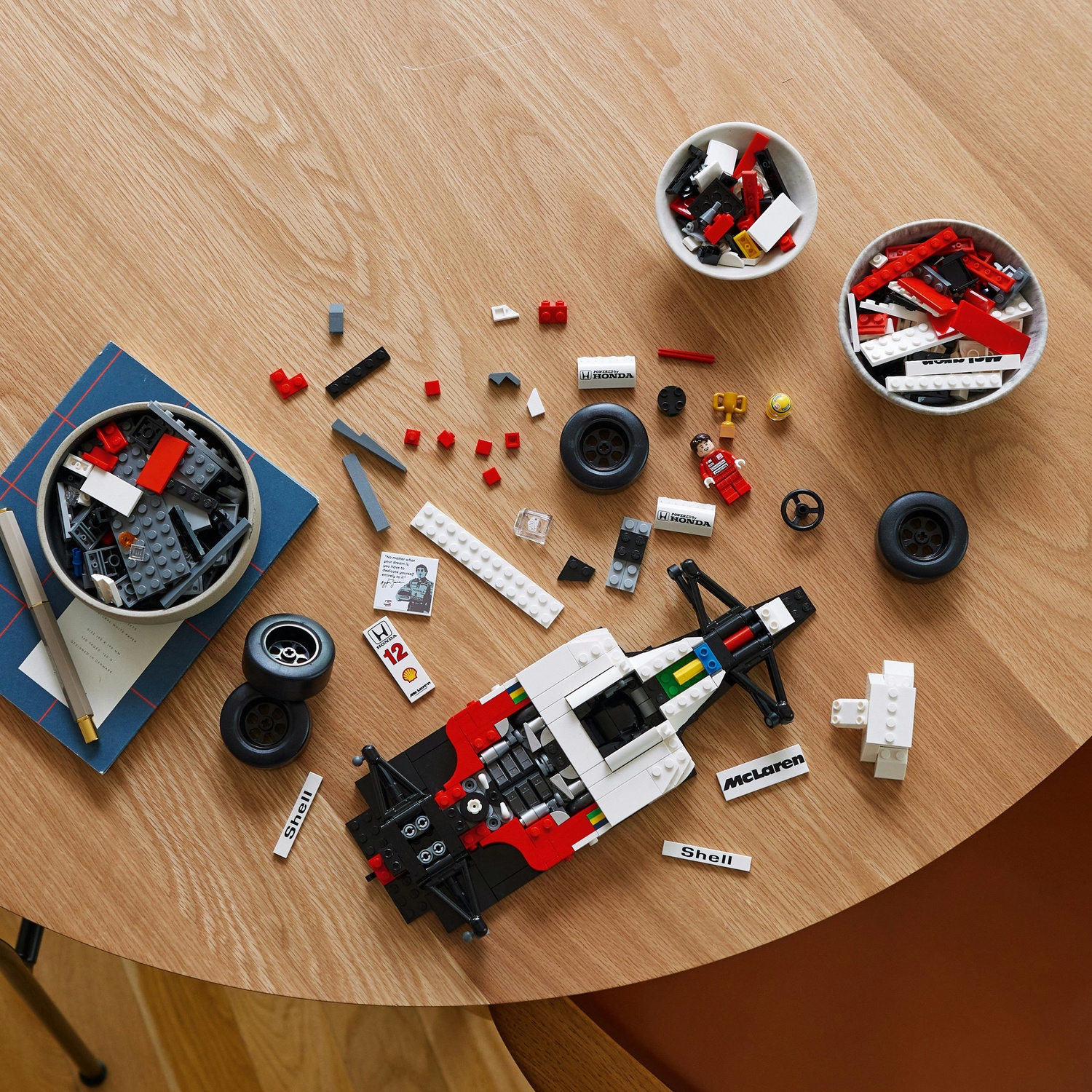 Un'auto da corsa Lego parzialmente assemblata su un tavolo di legno, circondata da componenti, ruote, mini-figure e ciotole di mattoncini Lego.