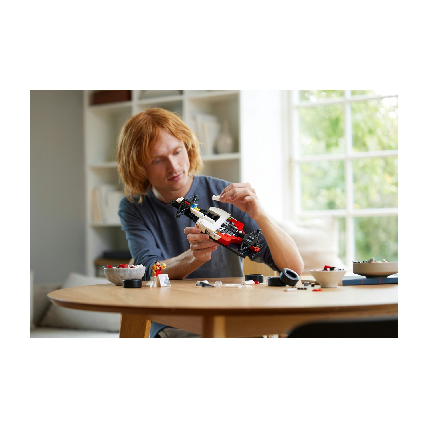 Un uomo dai capelli rossi assembla un'auto Lego su un tavolo di legno. Pezzi e pneumatici Lego sono sparsi sul tavolo.