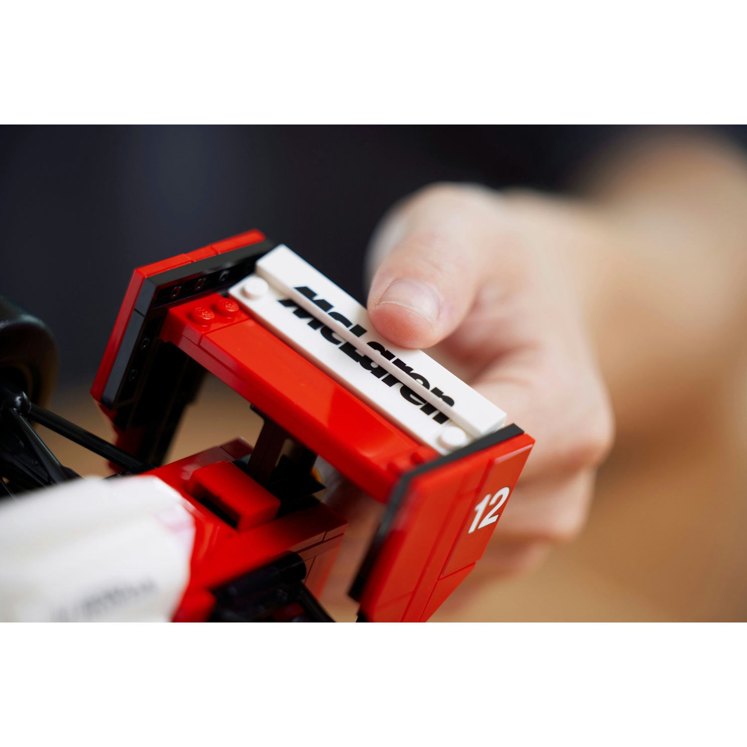 Una mano tiene un modello Lego McLaren. L'auto è rossa e bianca con il numero 12. La parola "McLaren" è visibile.