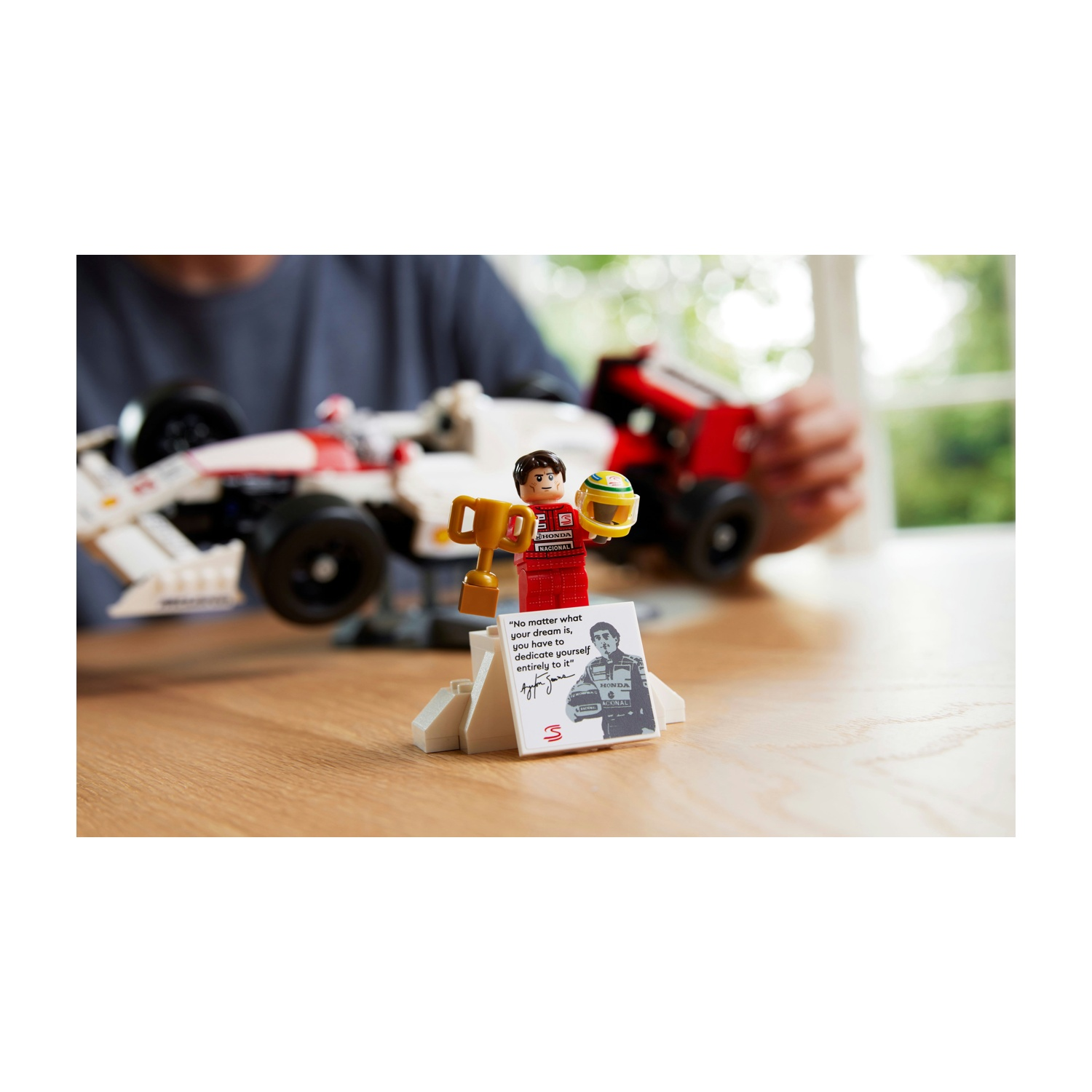 Un personaggio Lego tiene in mano un trofeo e un casco. Un modello di auto da corsa è sullo sfondo. Una citazione e una foto sono su una carta bianca.