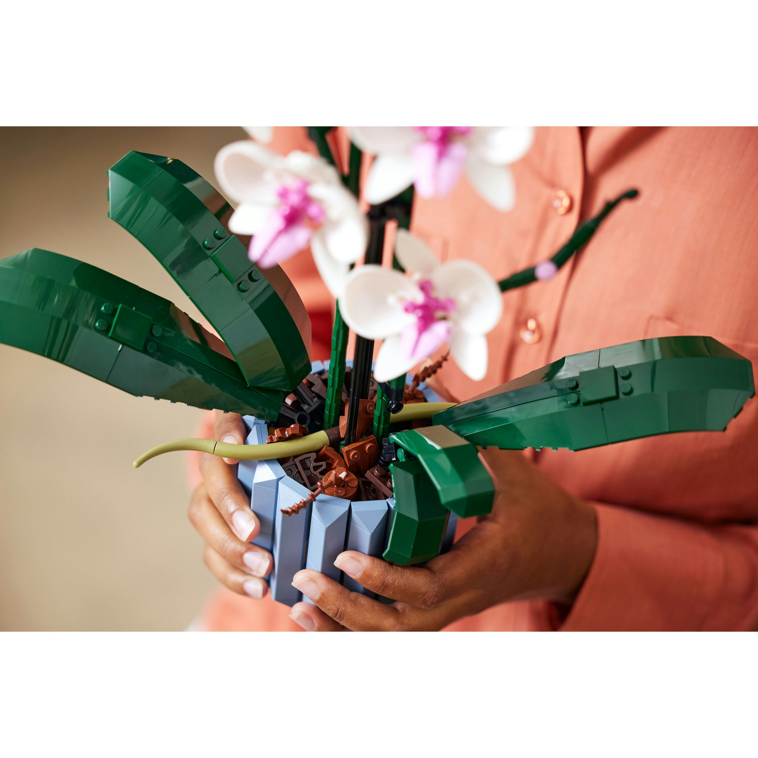 Una persona tiene in mano una pianta di orchidea Lego. La pianta ha fiori bianchi e rosa, foglie verdi ed è in un vaso blu.