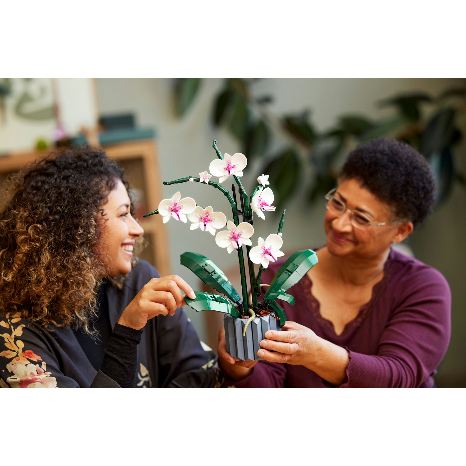 Due donne tengono una pianta di orchidea fatta con i Lego. L'orchidea ha fiori bianchi e rosa e foglie verdi.