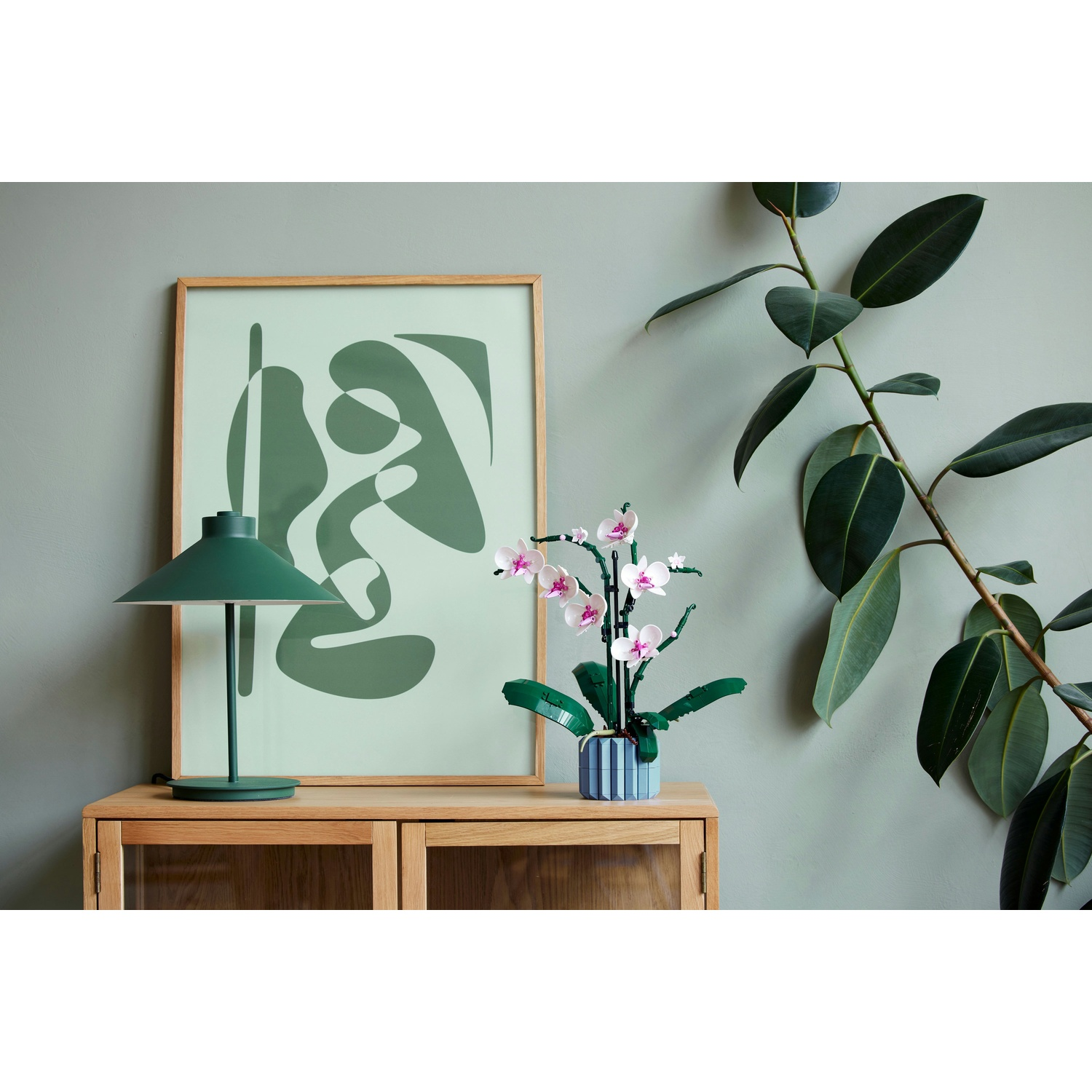 Un mobile in legno con lampada, quadro e orchidea. A destra, una grande pianta con foglie verdi.
