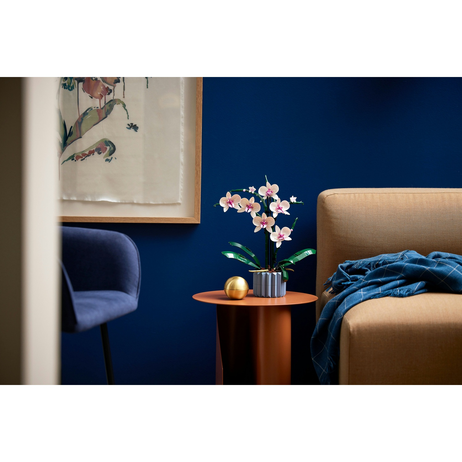 Un'orchidea in un vaso blu su un tavolino, accanto a un divano beige con una coperta blu. Un'opera d'arte incorniciata è appesa al muro.