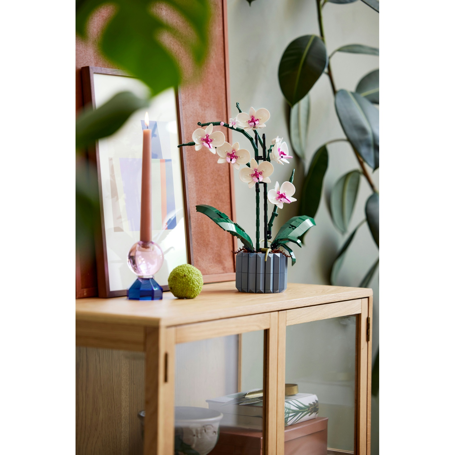 Un'orchidea in un vaso grigio su un mobile di legno, con una candela e un'arte incorniciata. Foglie verdi sullo sfondo.