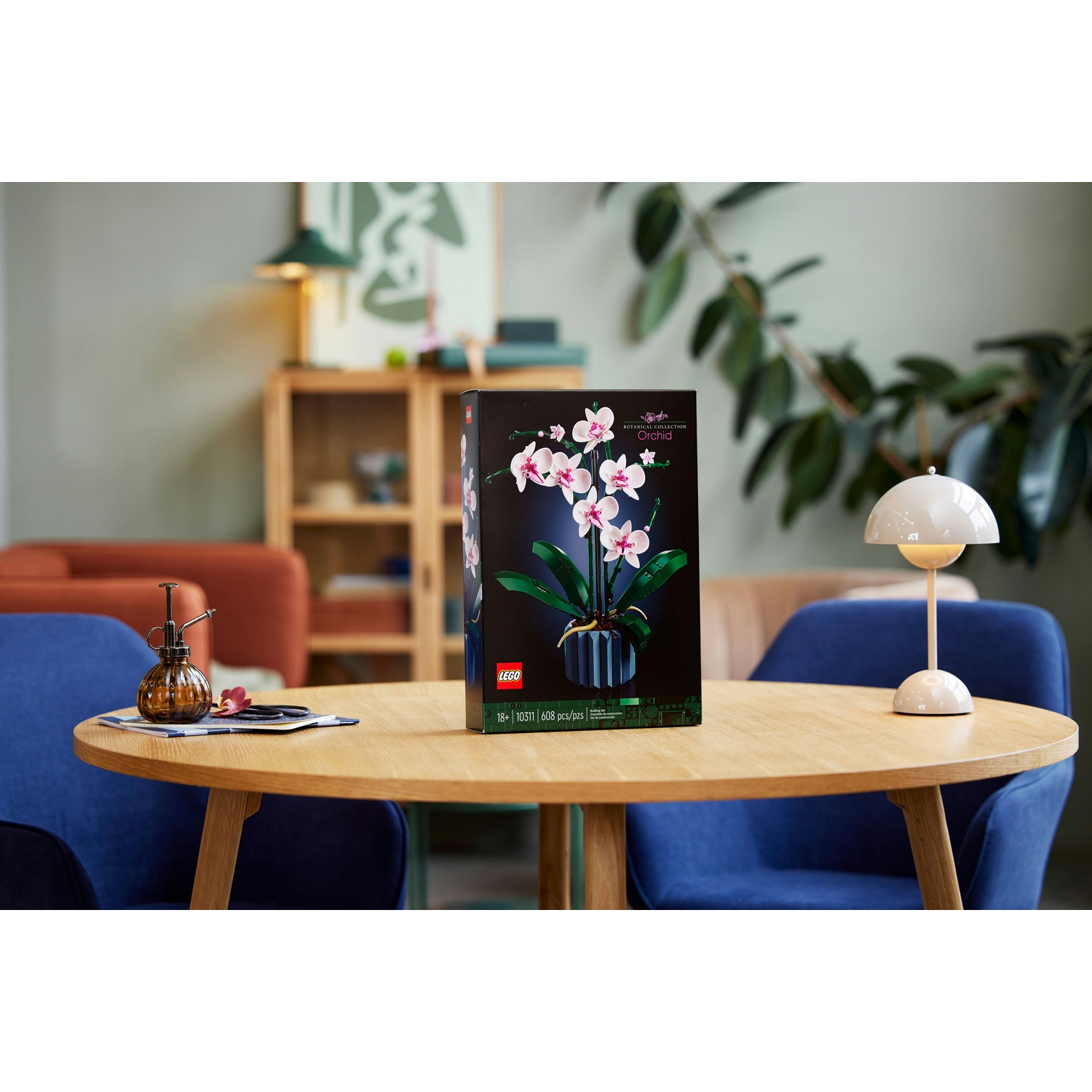 Una scatola del set LEGO Orchidea su un tavolo di legno, di fronte a sedie blu, una lampada e una pianta. Un piccolo spruzzatore è sul tavolo.