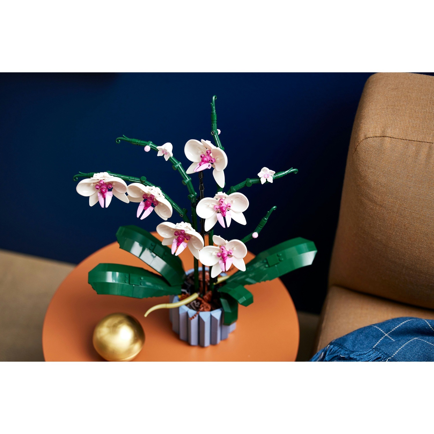 Un'orchidea Lego in un vaso su un tavolo arancione, una sfera dorata e una poltrona marrone sono vicini.