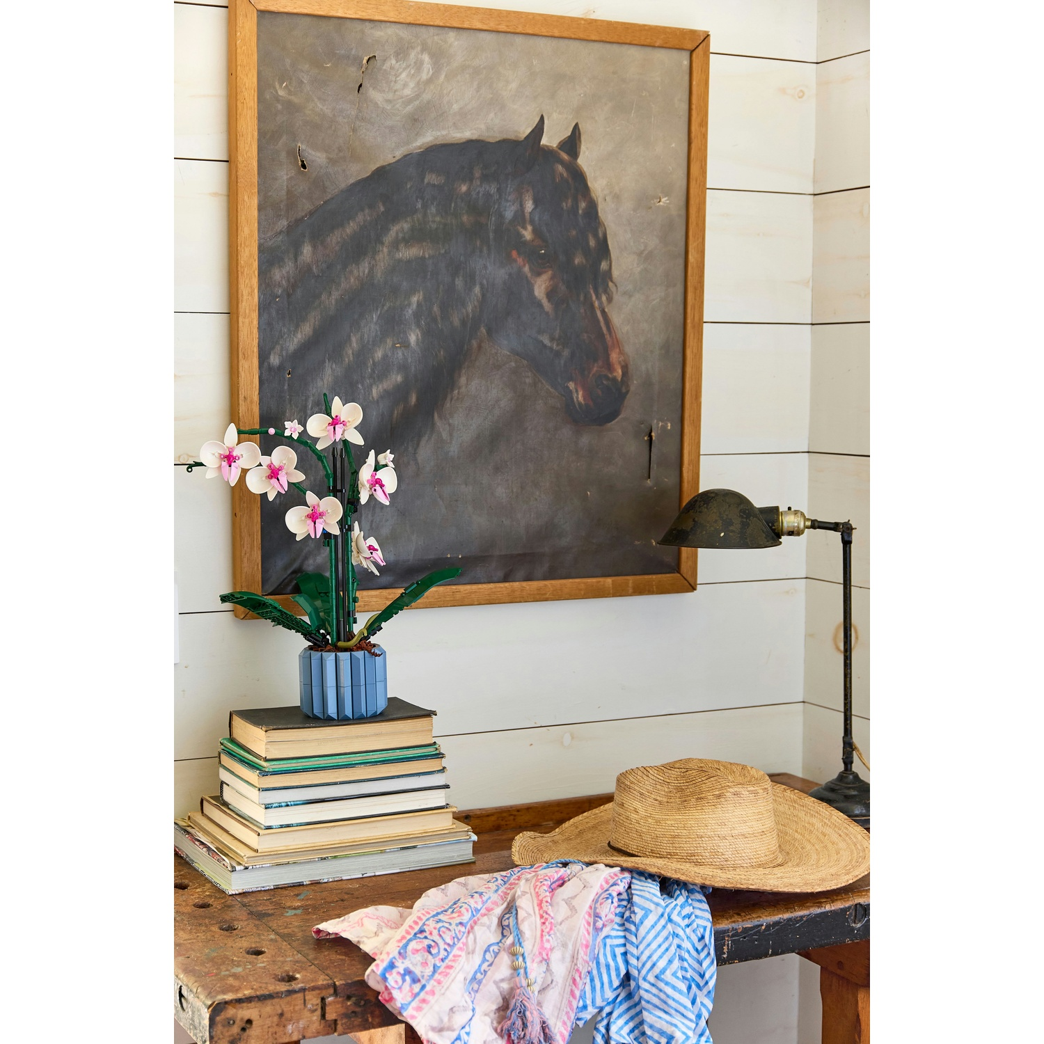 Una stanza con tavolo in legno, dipinto di cavallo, fiore, libri, cappello e lampada.
