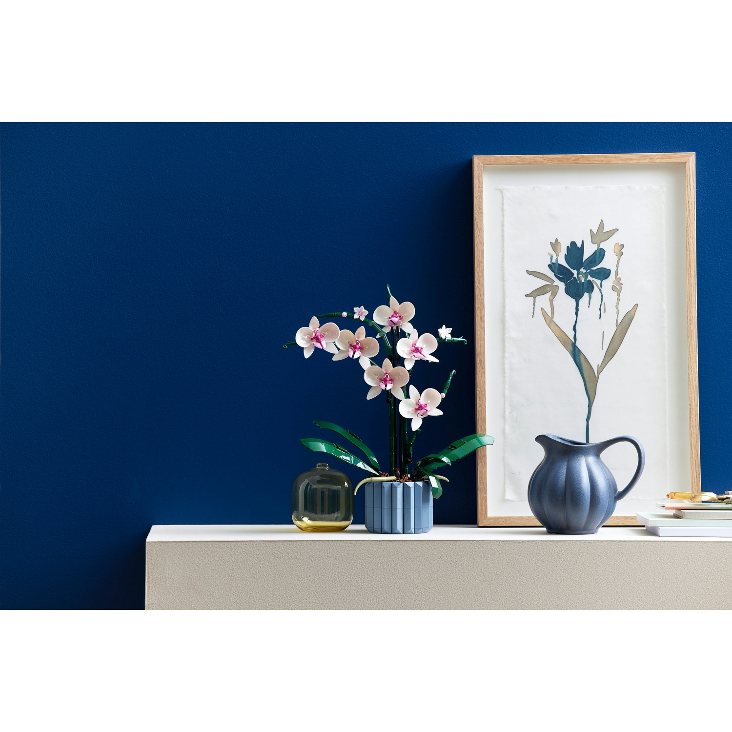 Un'orchidea in un vaso blu, un'opera d'arte incorniciata e una brocca blu sono su uno scaffale contro un muro blu scuro.