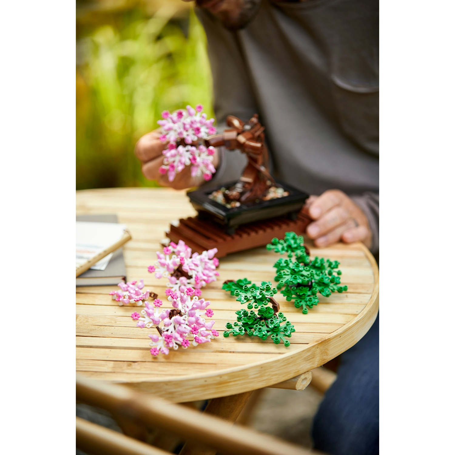 Una persona tiene in mano un fiore Lego rosa e bianco. Albero bonsai Lego e fogliame Lego verde su un tavolo di bambù. Sfondo verde sfocato.