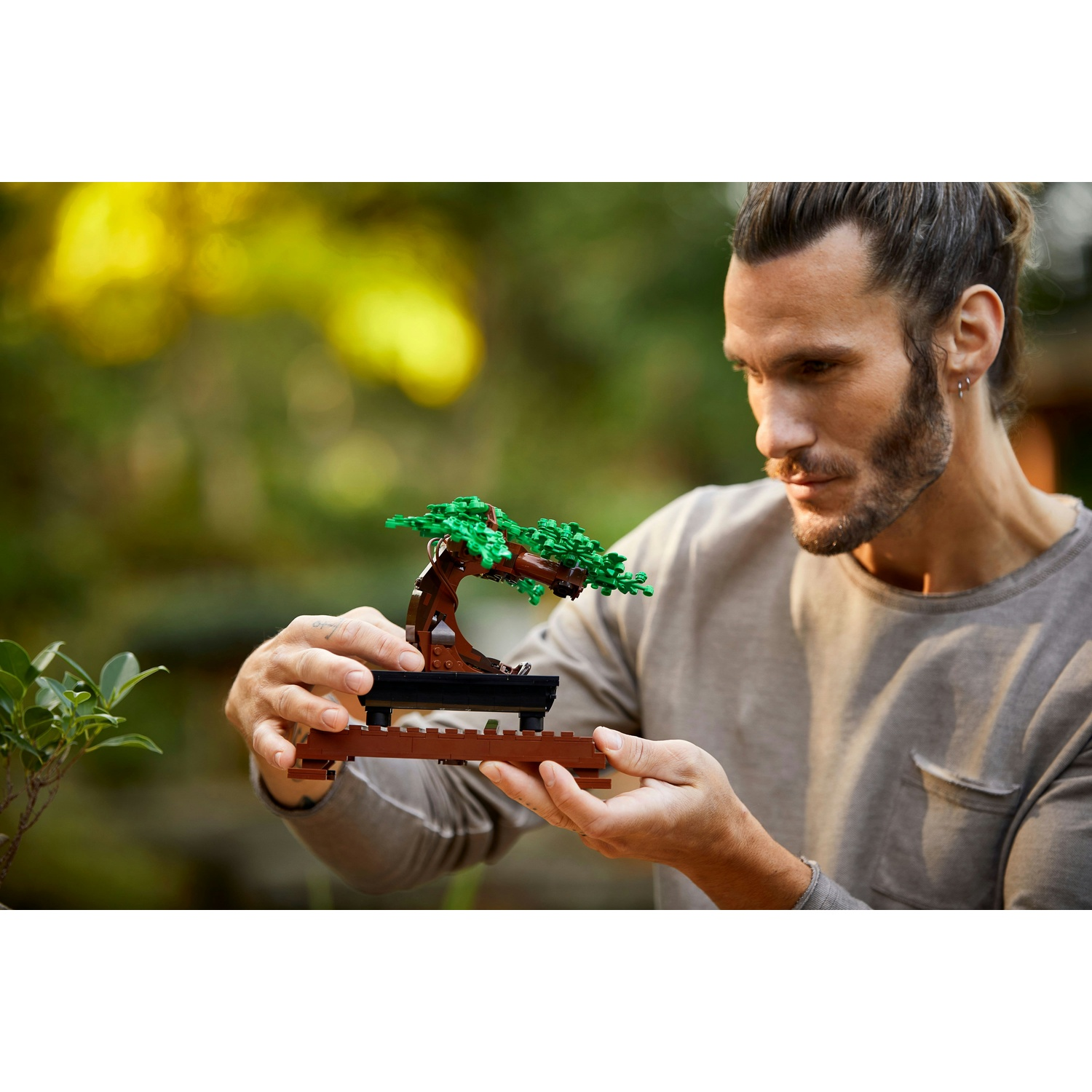 Un uomo tiene un bonsai Lego. L'albero è marrone e verde. L'uomo ha la barba e indossa una camicia grigia.