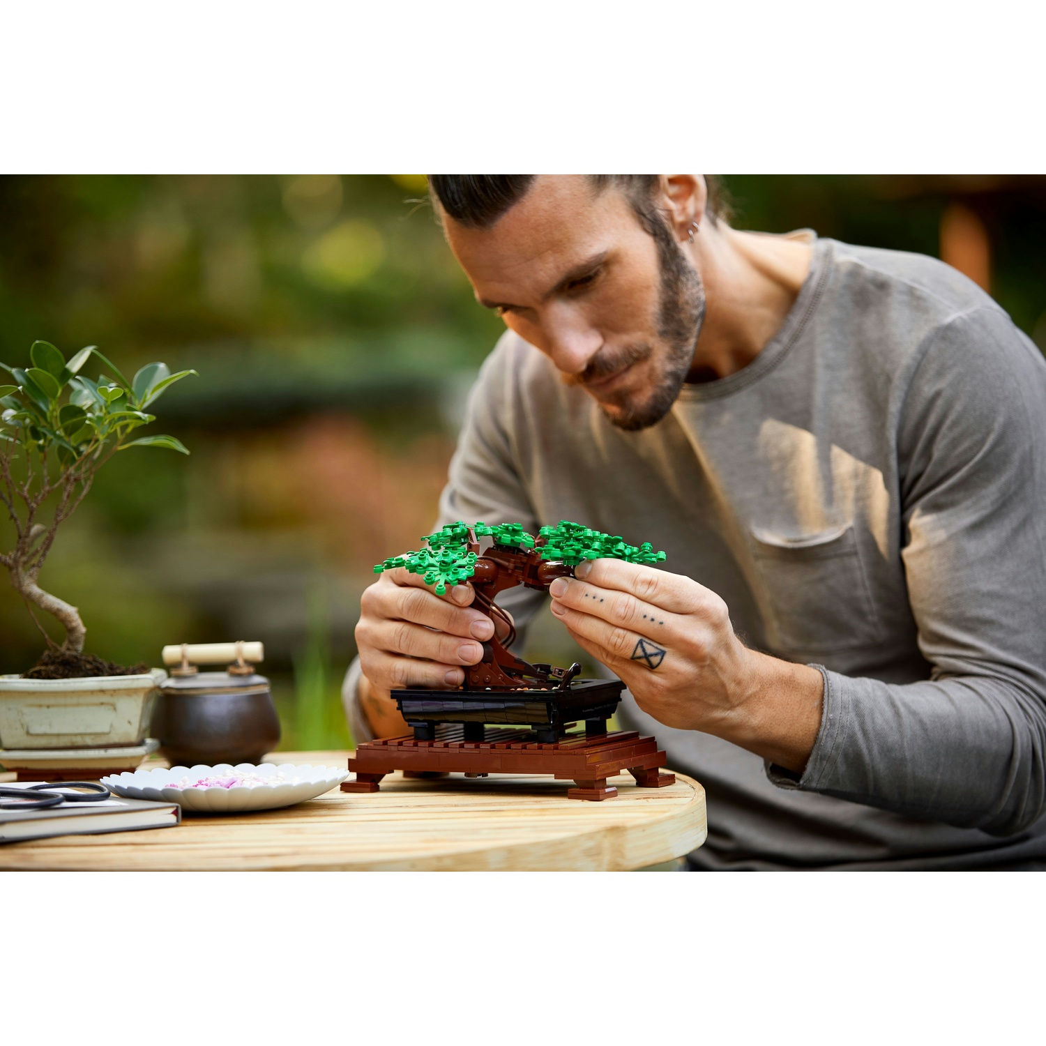 Un uomo assembla un bonsai Lego. Altri bonsai e attrezzi sono sul tavolo di legno. Lo sfondo è verde sfocato.