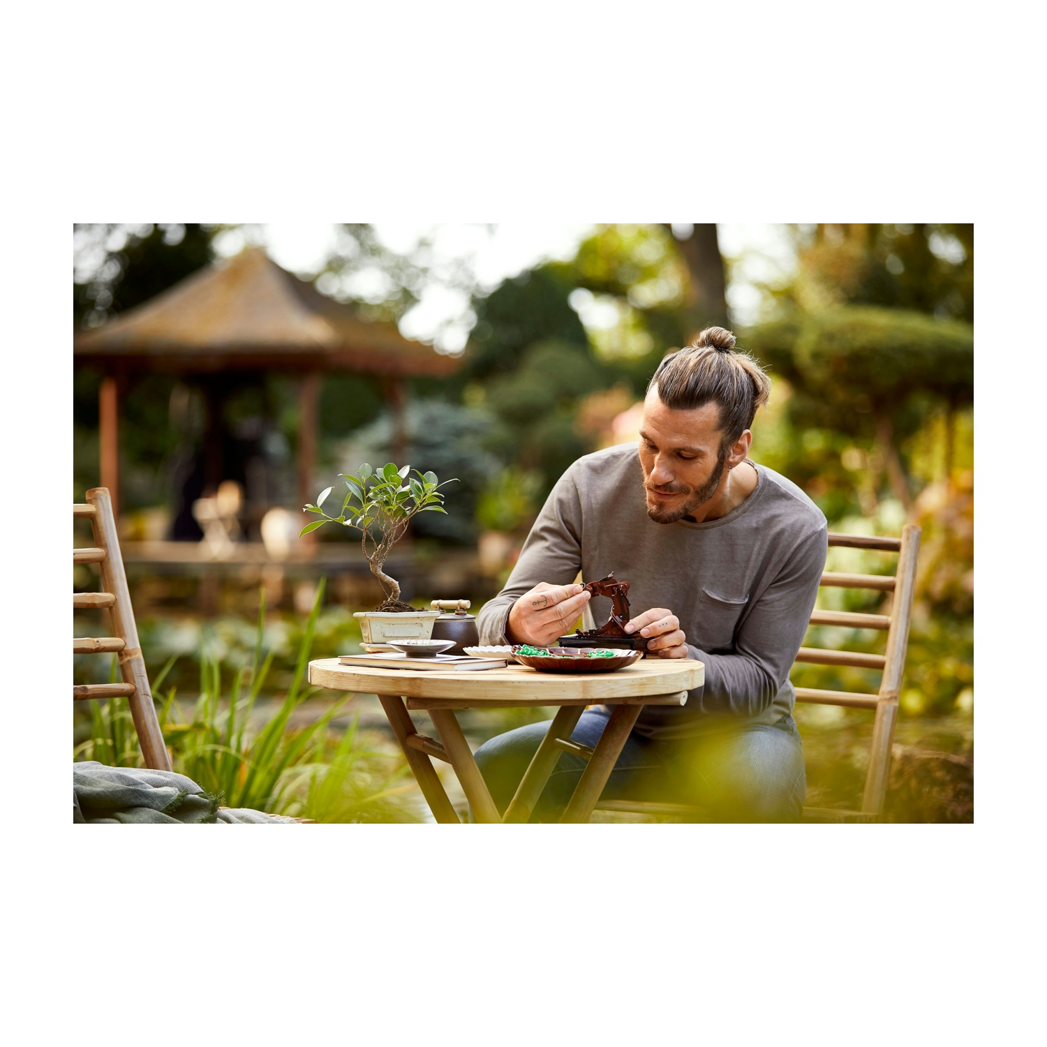 Un uomo con uno chignon lavora su un bonsai all'aperto. Un tavolino contiene attrezzi e piante. Un giardino e un gazebo sono sullo sfondo.
