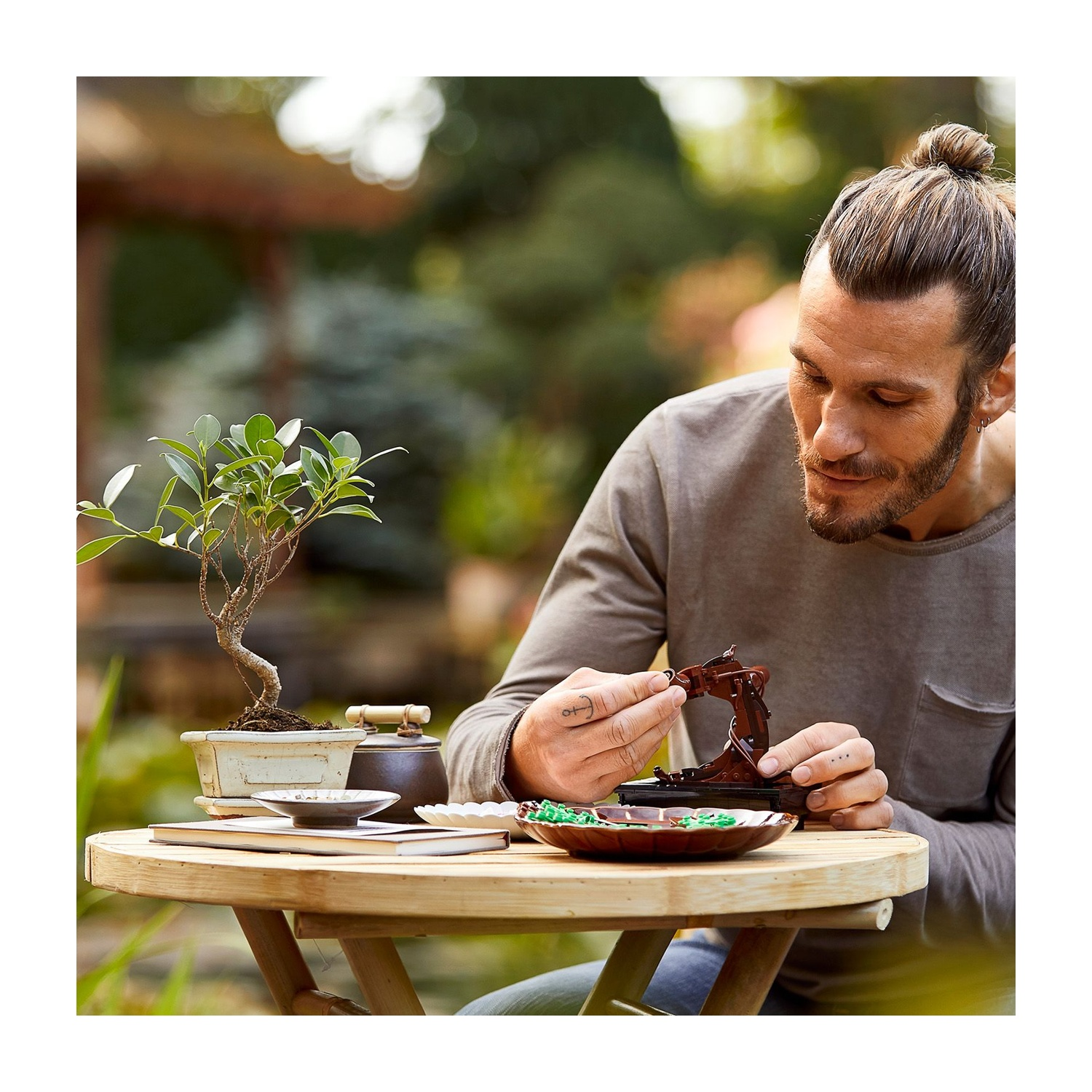Uomo con chignon costruisce struttura in miniatura su tavolo con bonsai, set da tè e piatto con pezzi verdi. Ambientazione esterna.