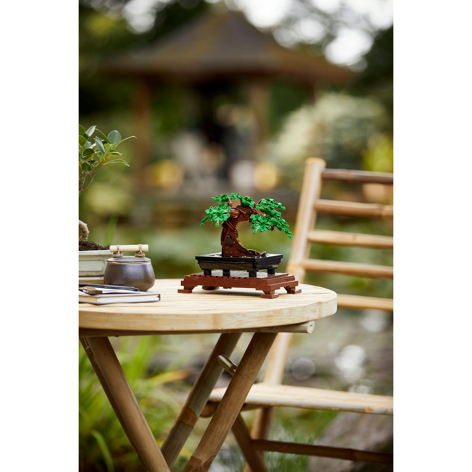 Un bonsai Lego è posizionato su un tavolo rotondo di legno, con una sedia e uno sfondo sfocato di un giardino con una pagoda.