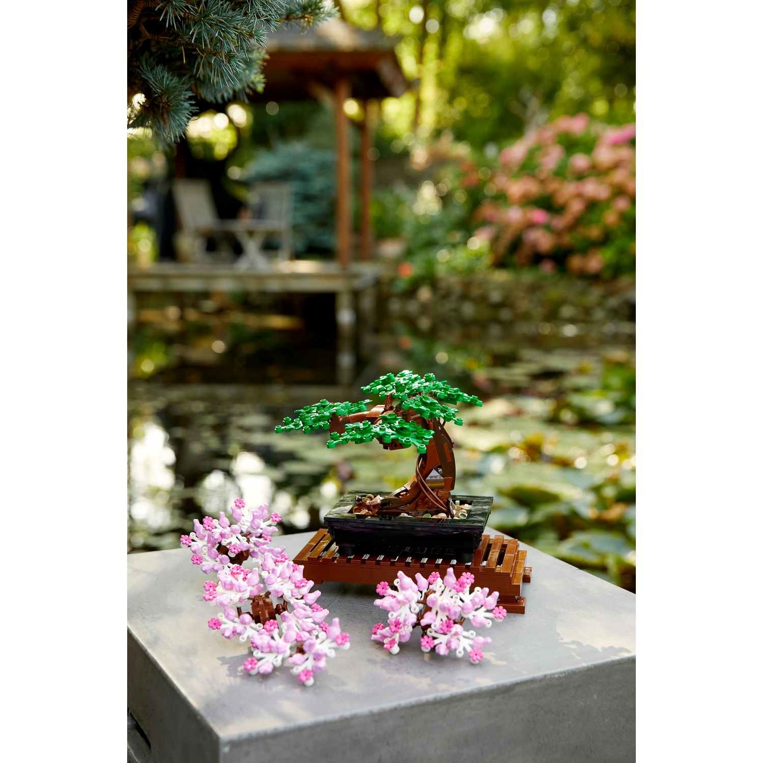 Un bonsai Lego con foglie verdi e tronco marrone è su un supporto di legno, con fiori rosa. Un giardino sfocato sullo sfondo.