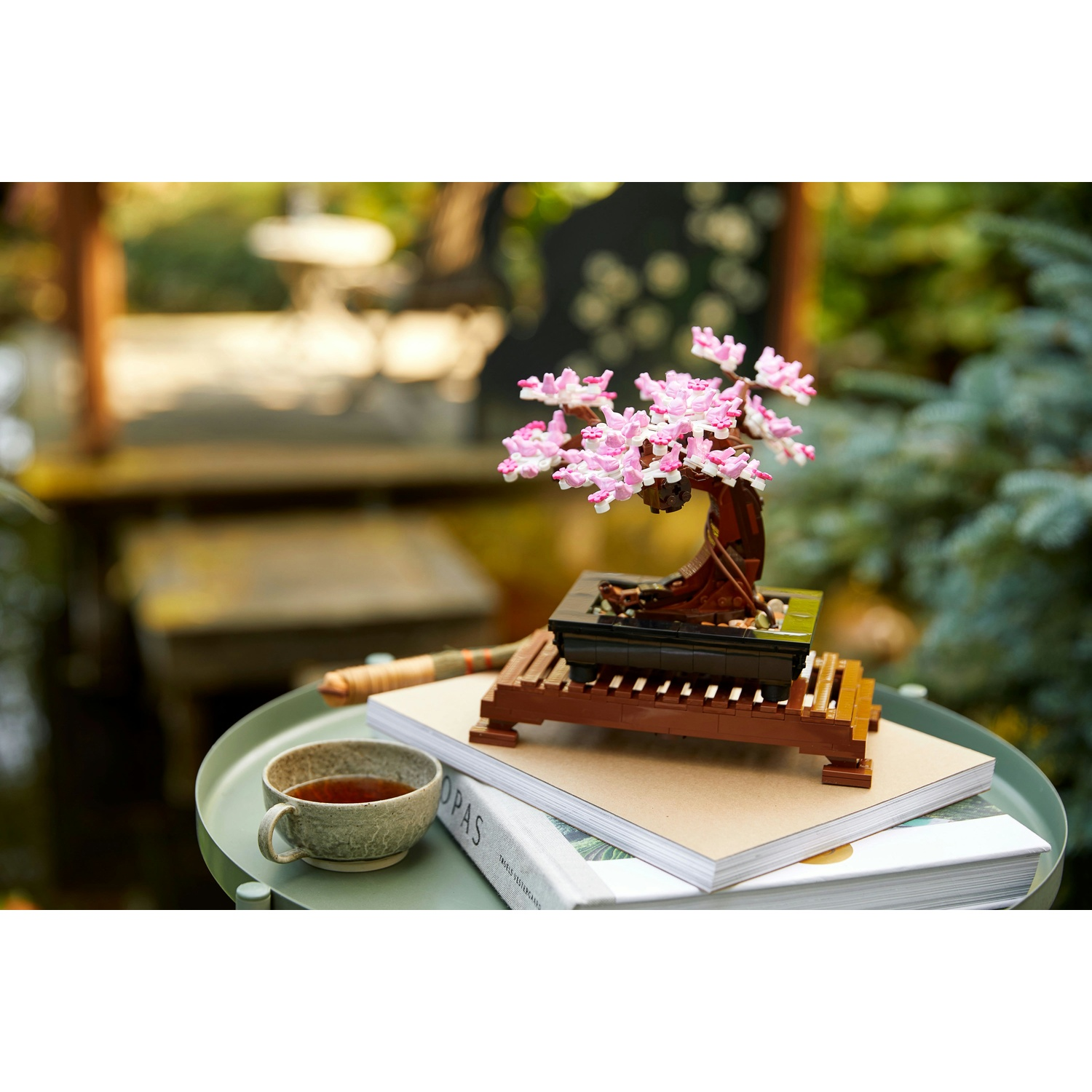 Un bonsai con fiori rosa è su libri. Una tazza di tè è su un tavolo. Lo sfondo è vegetazione sfocata.
