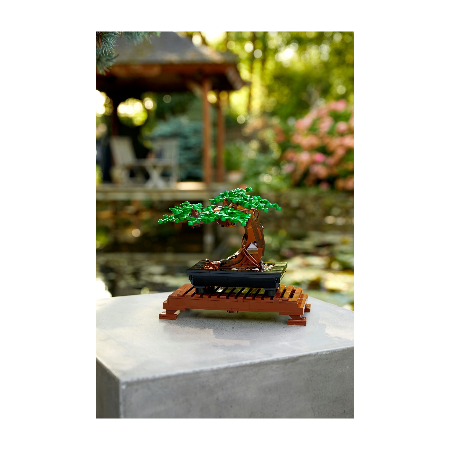 Un bonsai Lego su un supporto marrone è posto su una superficie di cemento. Sullo sfondo, un gazebo e fogliame verde.