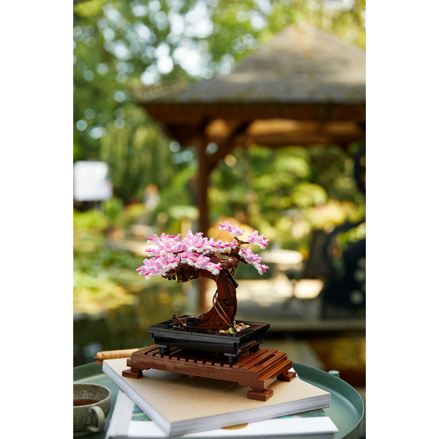 Un bonsai Lego con fiori rosa su un tavolo con libri. Sullo sfondo un gazebo e vegetazione.