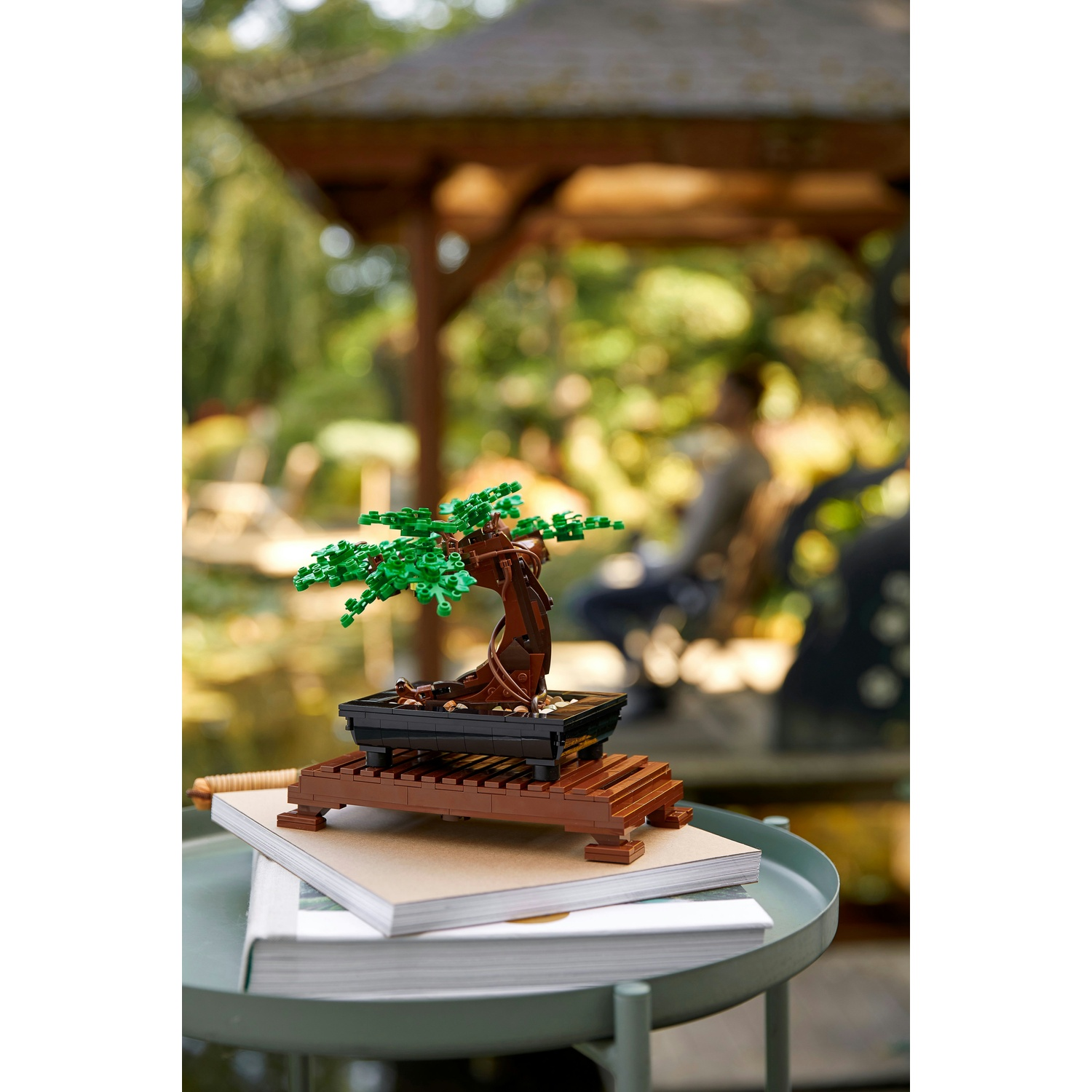 Un bonsai Lego con foglie verdi e tronco marrone è su un tavolo con libri. Uno sfondo sfocato mostra un gazebo e alberi.