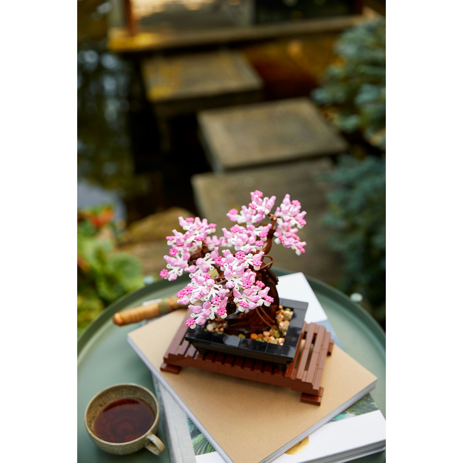 Un albero bonsai Lego con fiori rosa e bianchi si trova su libri e un tavolo, con una tazza di tè in primo piano e un giardino sullo sfondo.