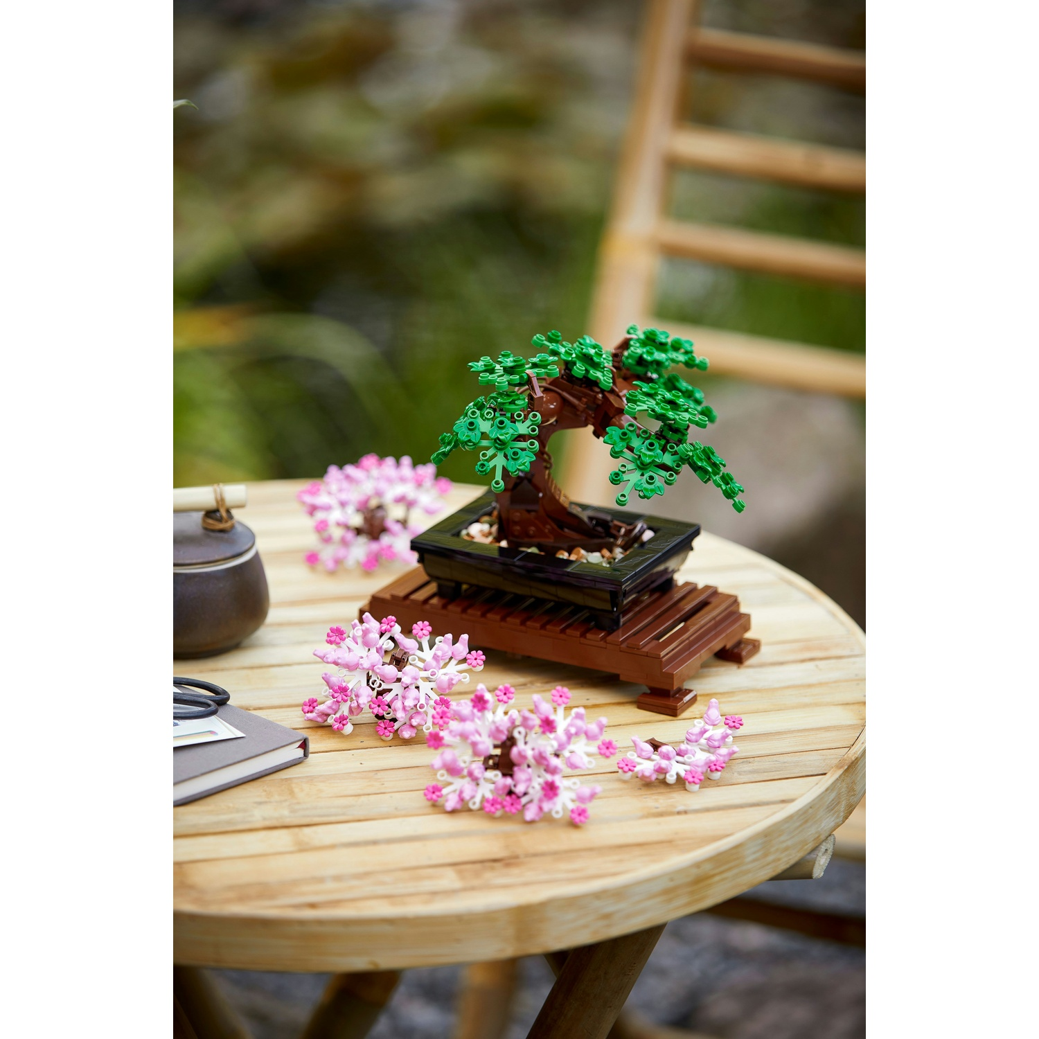 Un bonsai Lego con foglie verdi e tronco marrone è su un tavolo di legno con fiori rosa. Sono presenti anche un set da tè e un taccuino.