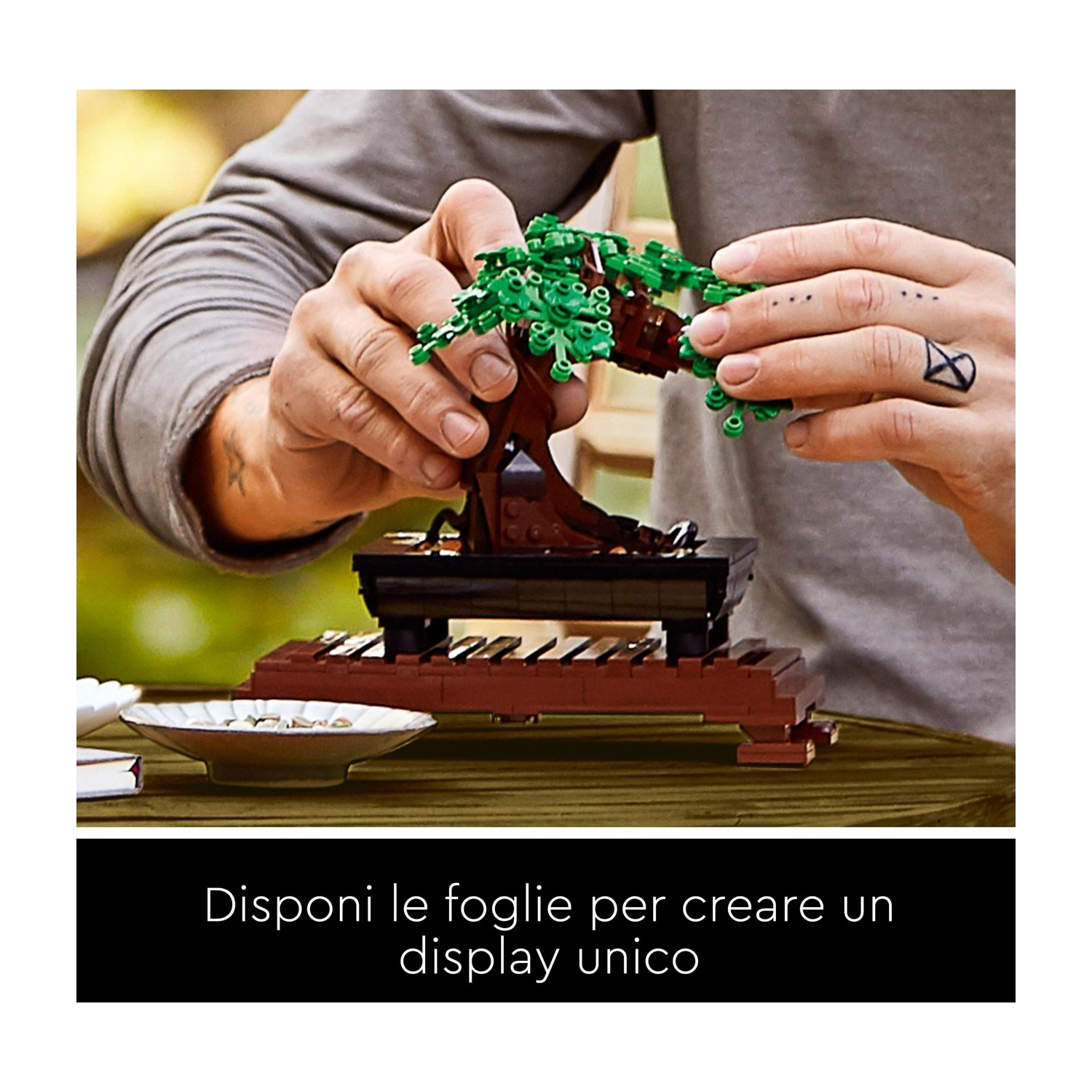 Mani che assemblano un bonsai Lego. Il tronco marrone è su un vaso nero, e sono attaccate foglie verdi. Un tatuaggio è visibile.