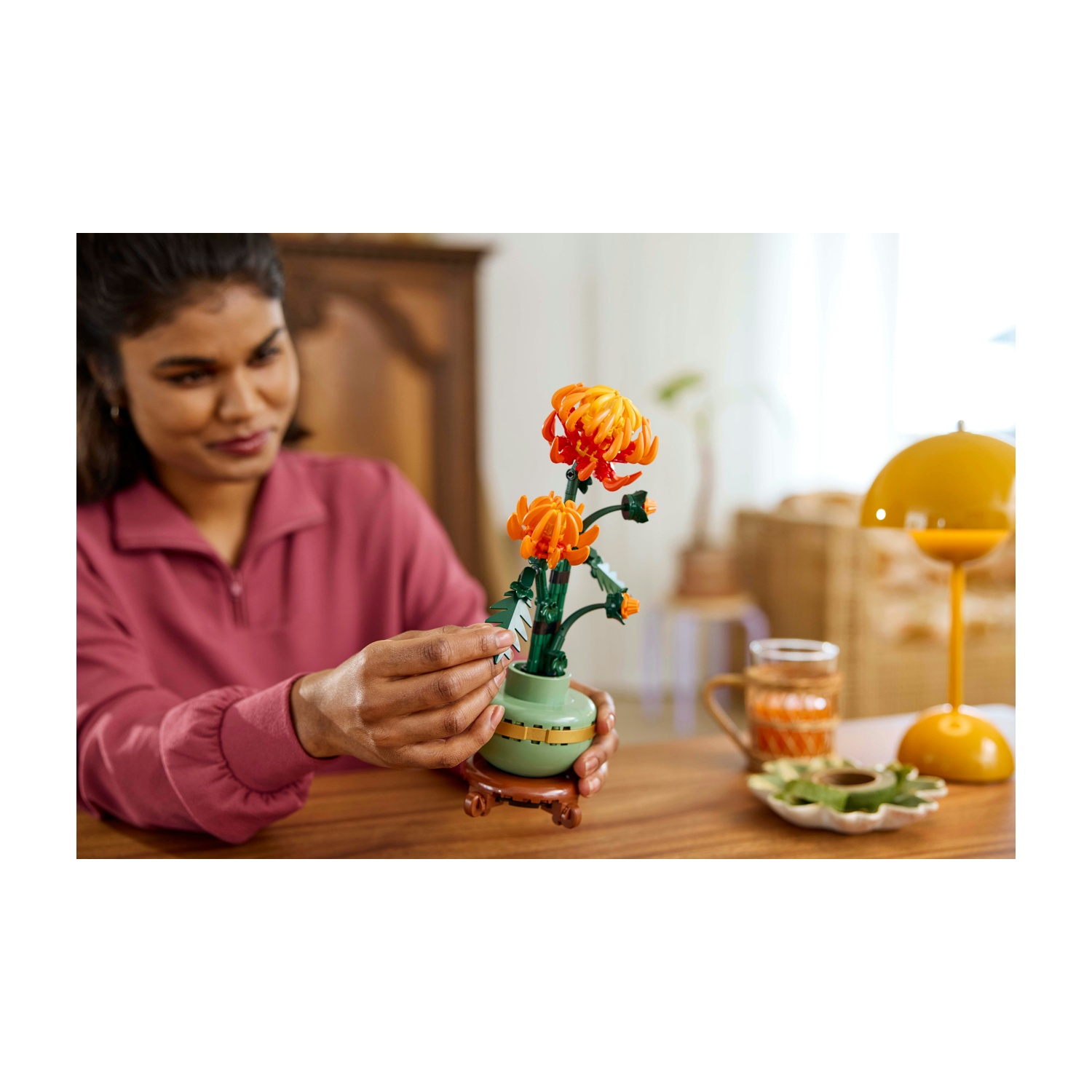 Donna che sistema fiori arancioni in un vaso verde su un tavolo. Sul tavolo ci sono anche una lampada gialla e una bevanda.