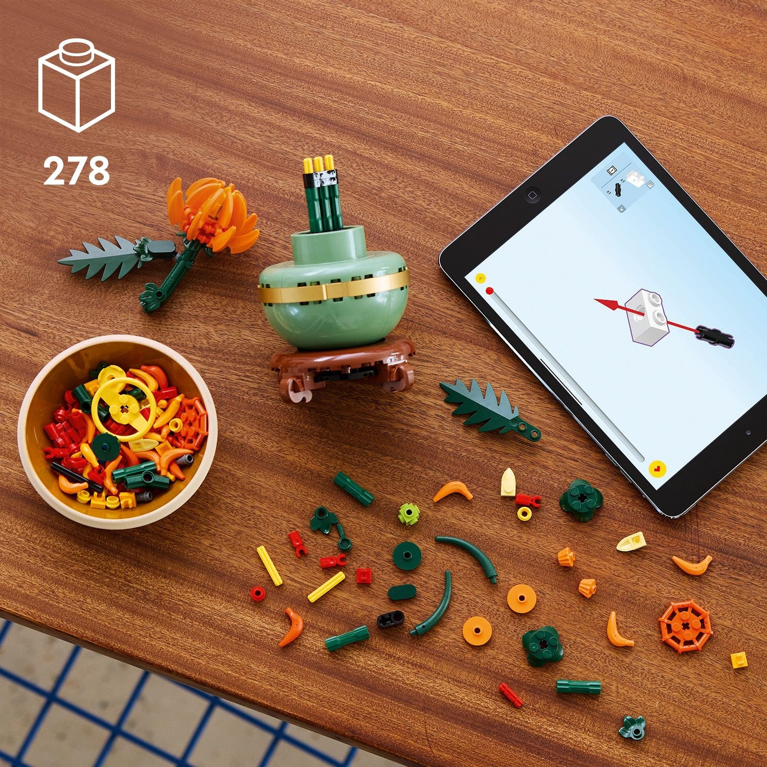 Set Lego con vari pezzi colorati, una ciotola, un fiore, un vaso verde e un tablet con le istruzioni su un tavolo di legno.